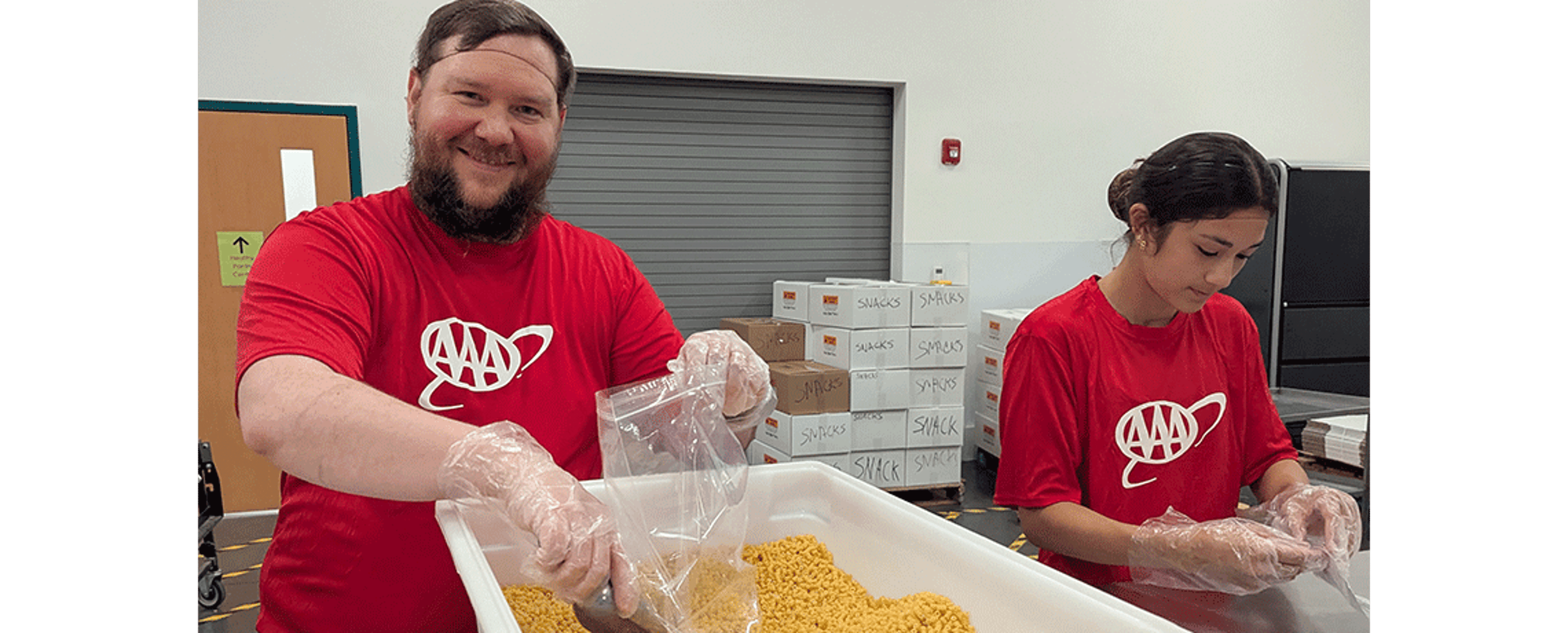 A bearded man in a red AAA shirt smiles while scooping dry pasta into a plastic bag at a volunteer station. A young woman next to him works on sealing bags. Boxes labeled “snacks” are stacked in the background