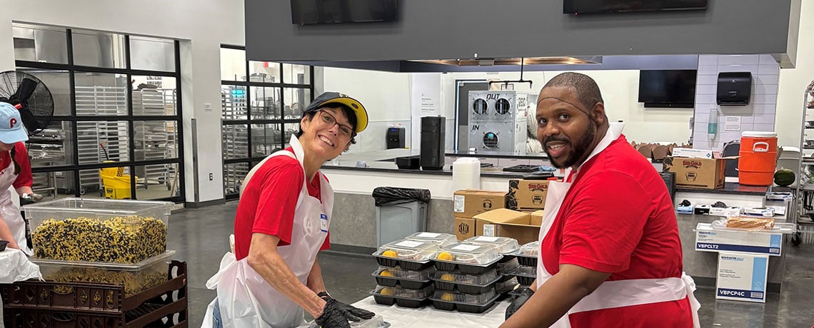 Two volunteers wearing red shirts, white aprons, and gloves stand behind a table of pre-packaged meals in a commercial kitchen labeled “The Wawa Demonstration Kitchen.” Both are smiling and working at a long table covered in meal trays.
