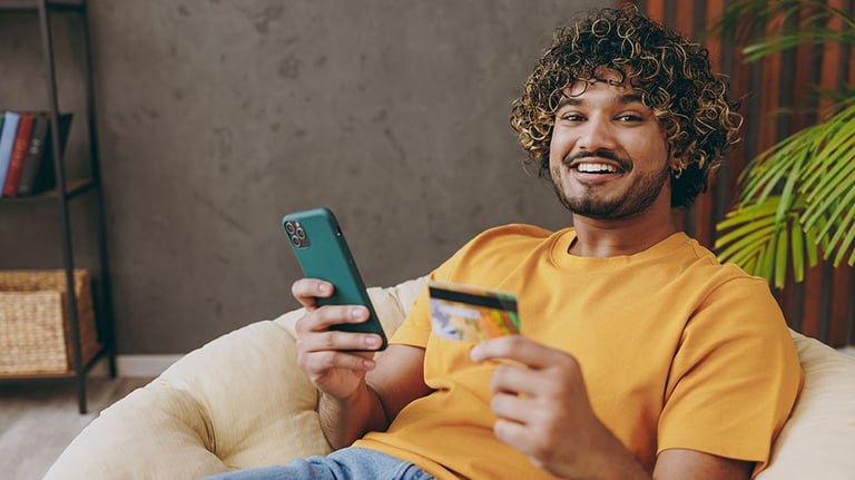 Young Indian man wear casual clothes hold mobile cell phone credit bank card shopping online sits in armchair stay at home hotel flat rest relax spend free spare time in living room. Lounge concept. By ViDi Studio