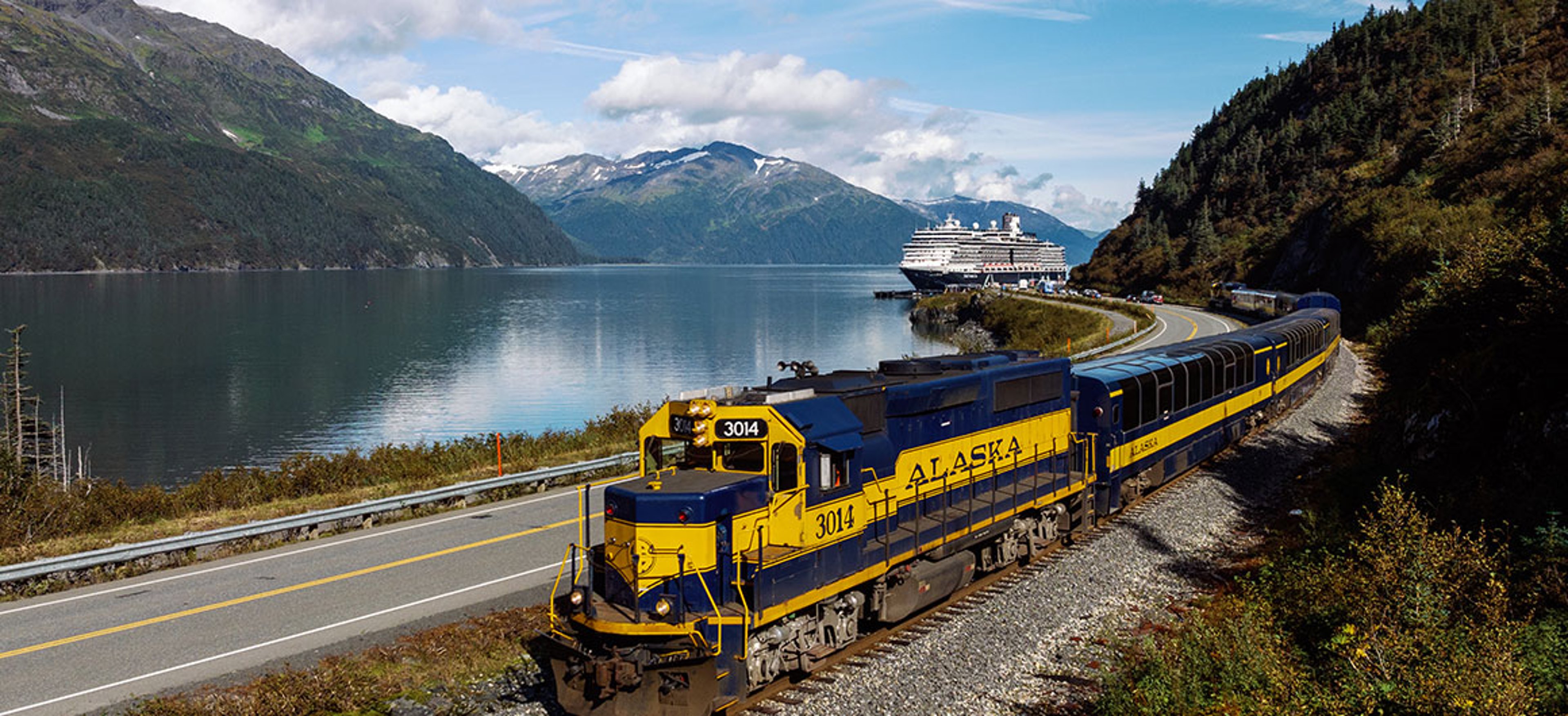 Cruise ship docked beside a passenger train at the port of Whittier, Alaska, with mountains in the background.