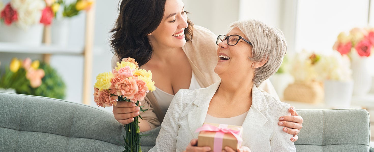 mother holding gift and daughter holding flowers looking at each other smiling