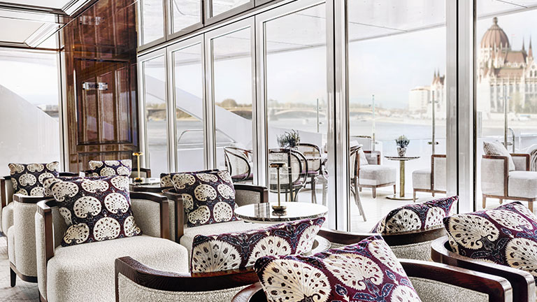Cushioned lounge chairs beside floor-to-ceiling windows overlooking the Danube River and Budapest.