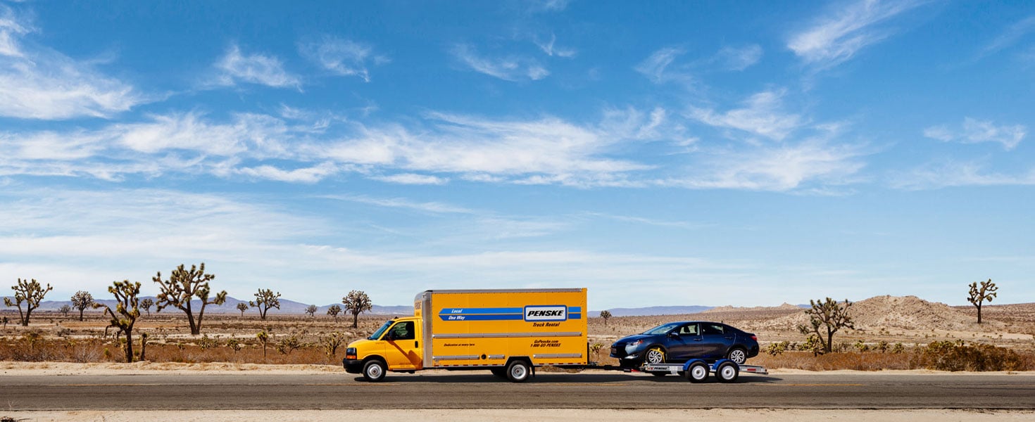 Penske rental truck hauling a car while driving through an open area with a bright blue sky