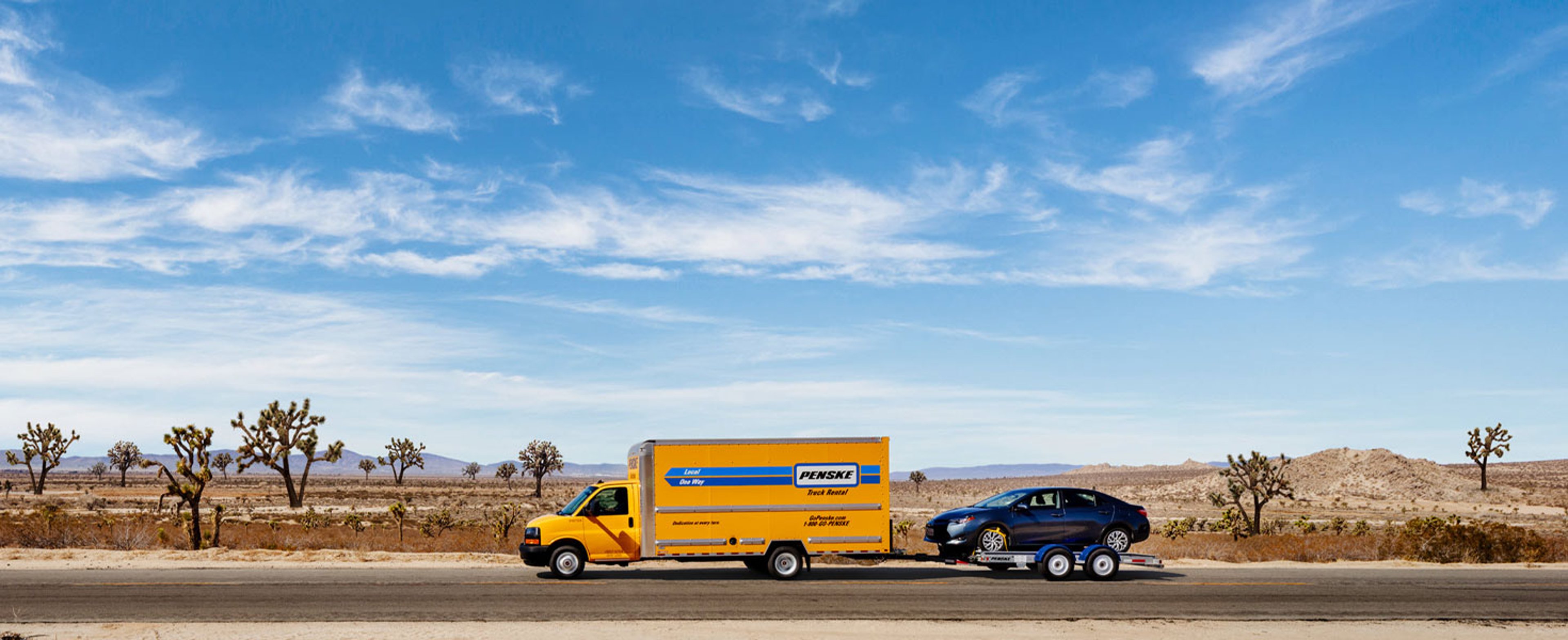 Penske rental truck hauling a car while driving through an open area with a bright blue sky