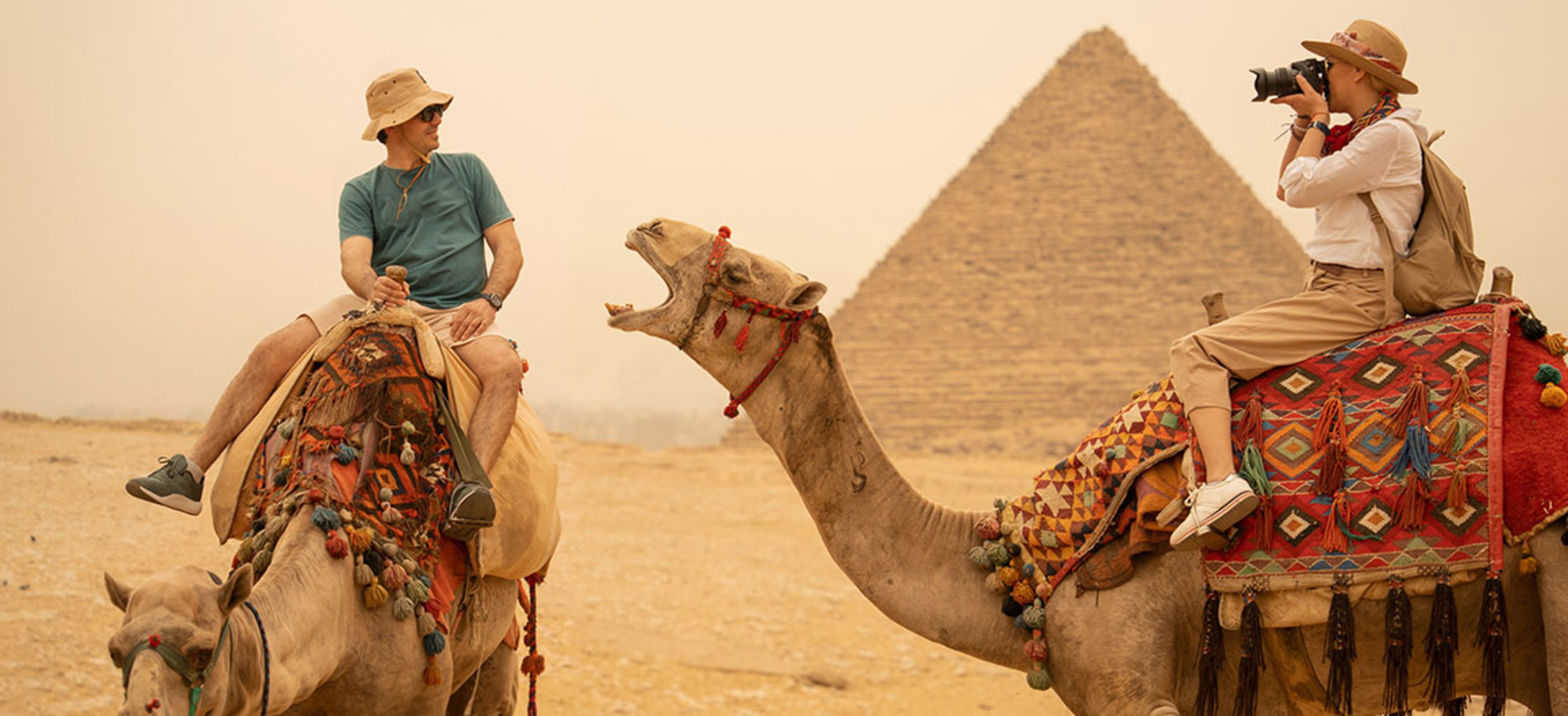 Adventurous Tourists Riding Camels by the Pyramids of Giza on a Dusty Day in Egypt; photo by valentinrussanov/iStock.com