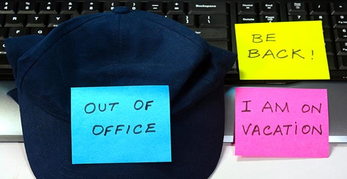 A baseball cap with sticky notes reading "OUT OF OFFICE," "BE BACK!" and "I AM ON VACATION" on a laptop keyboard.
