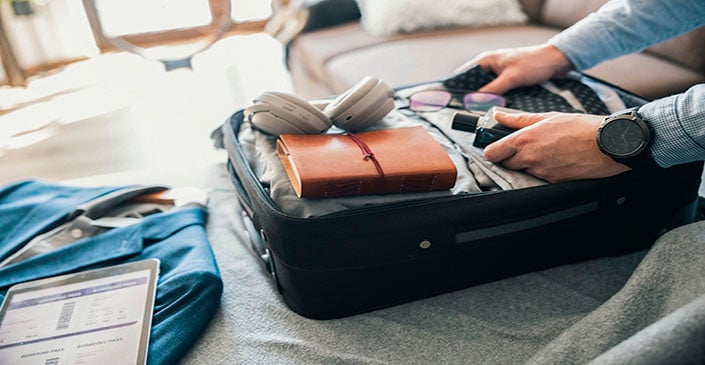 A person packing an open suitcase with headphones, notebook, remote, and clothing.