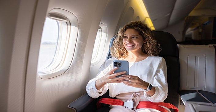 Person smiling while sitting in an airplane seat, holding a smartphone, with a window showing daylight outside.