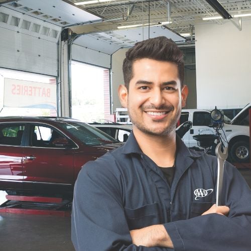 A smiling mechanic in a blue uniform holds a wrench in an auto repair shop with cars in the background.
