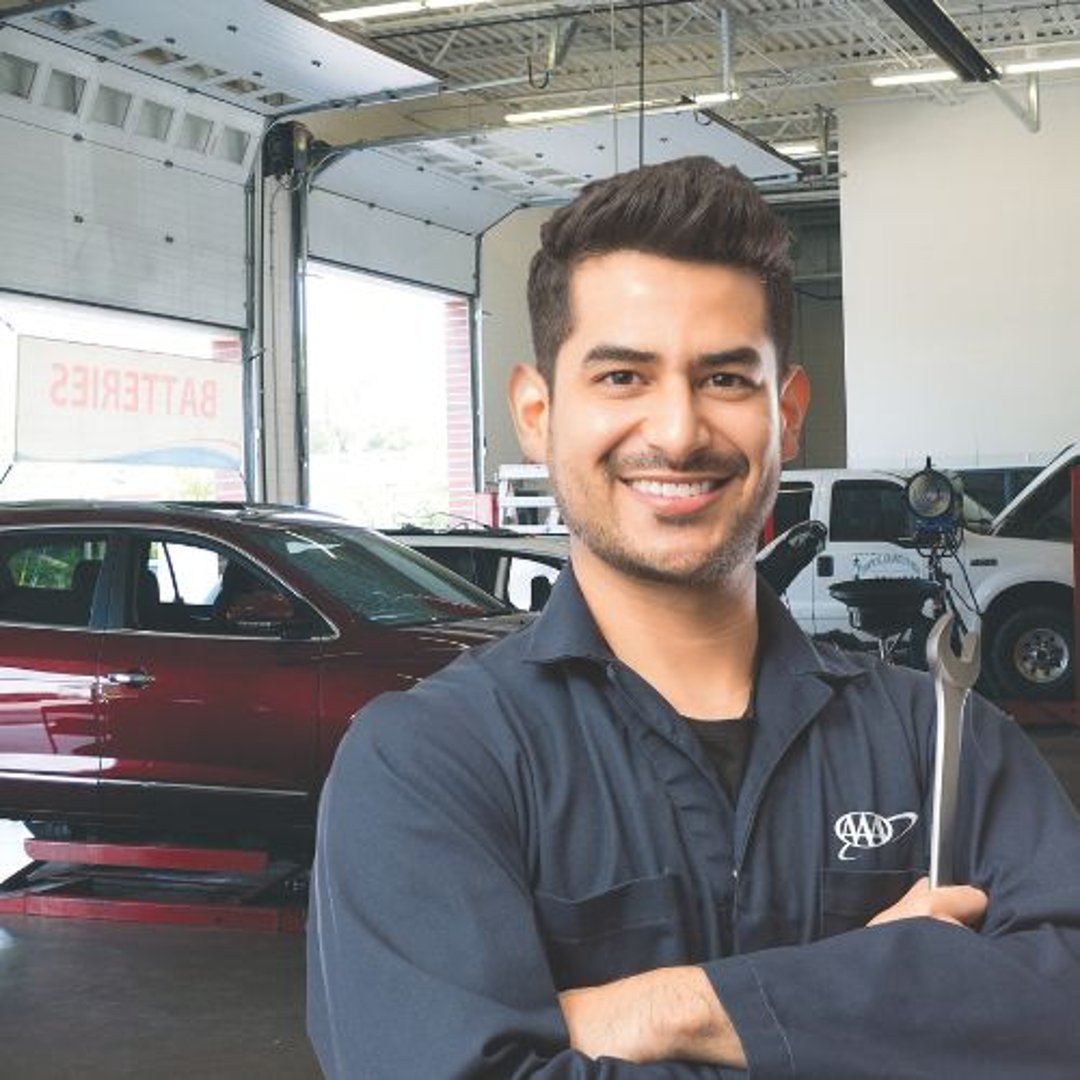 A smiling mechanic in a blue uniform holds a wrench in an auto repair shop with cars in the background.