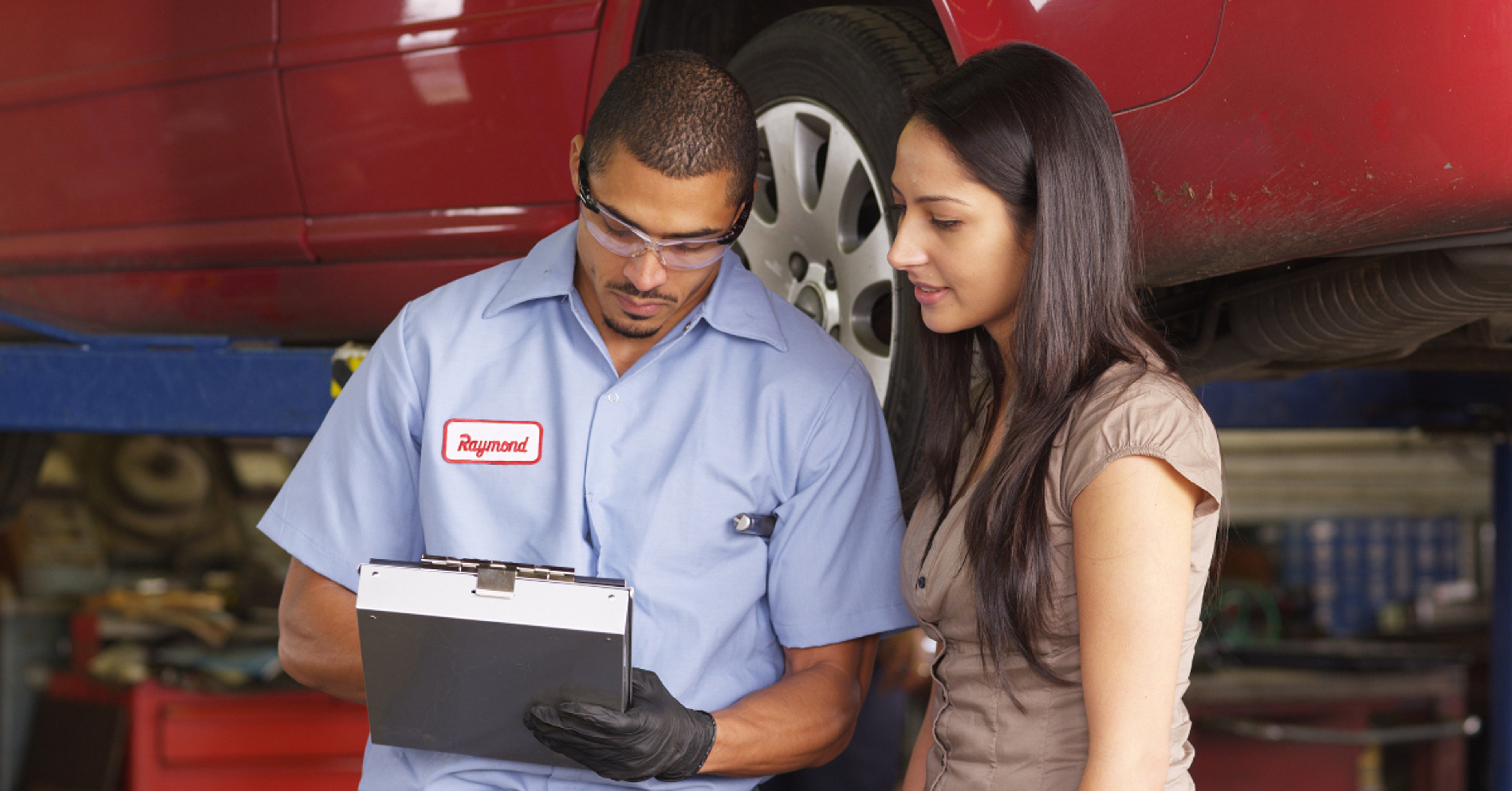 Car care technician explaining auto repair work to customer, with red sedan on jack stands in background 