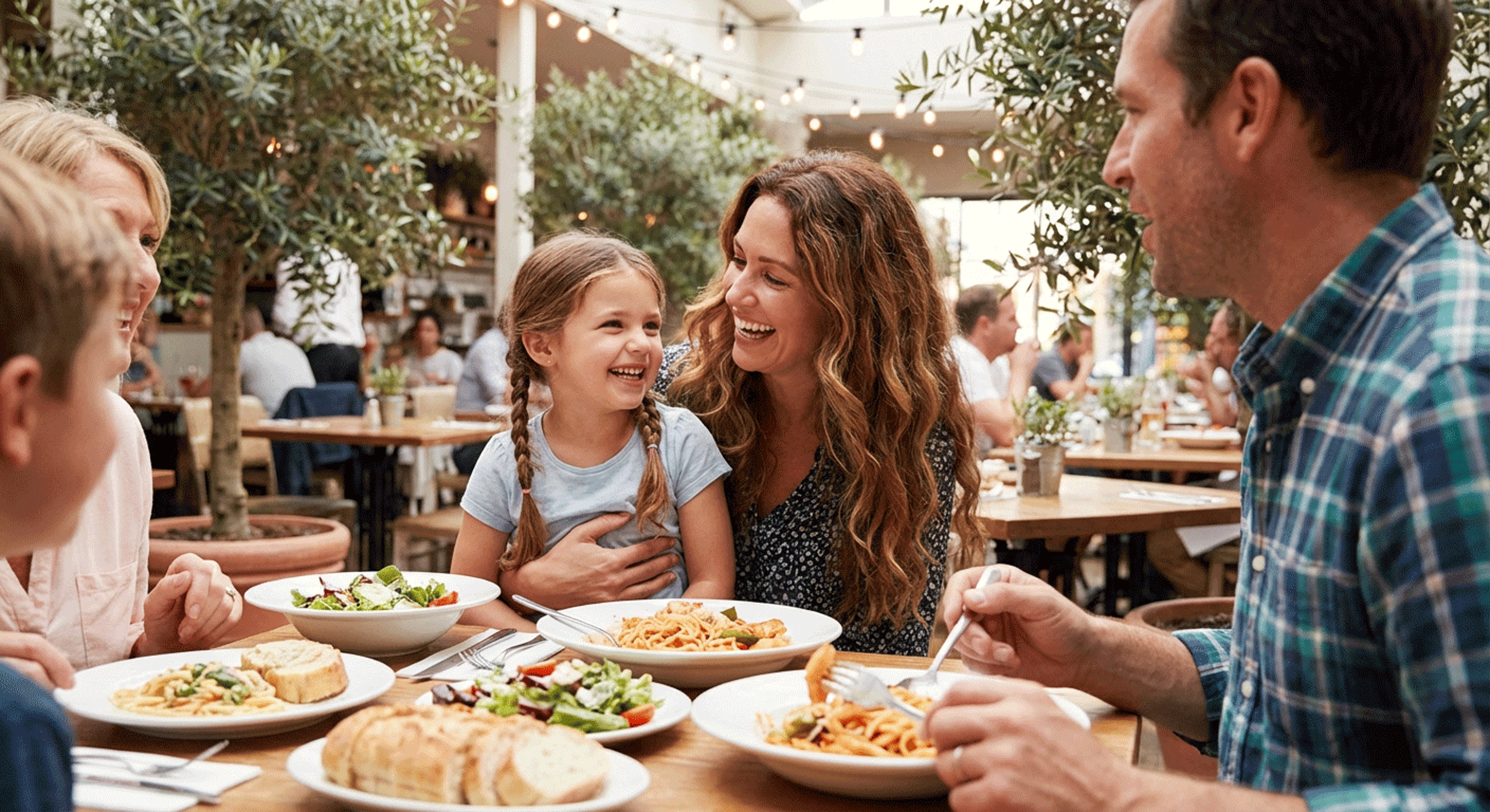 Family enjoying a meal in a bright restaurant with plates of pasta and salad on the table.