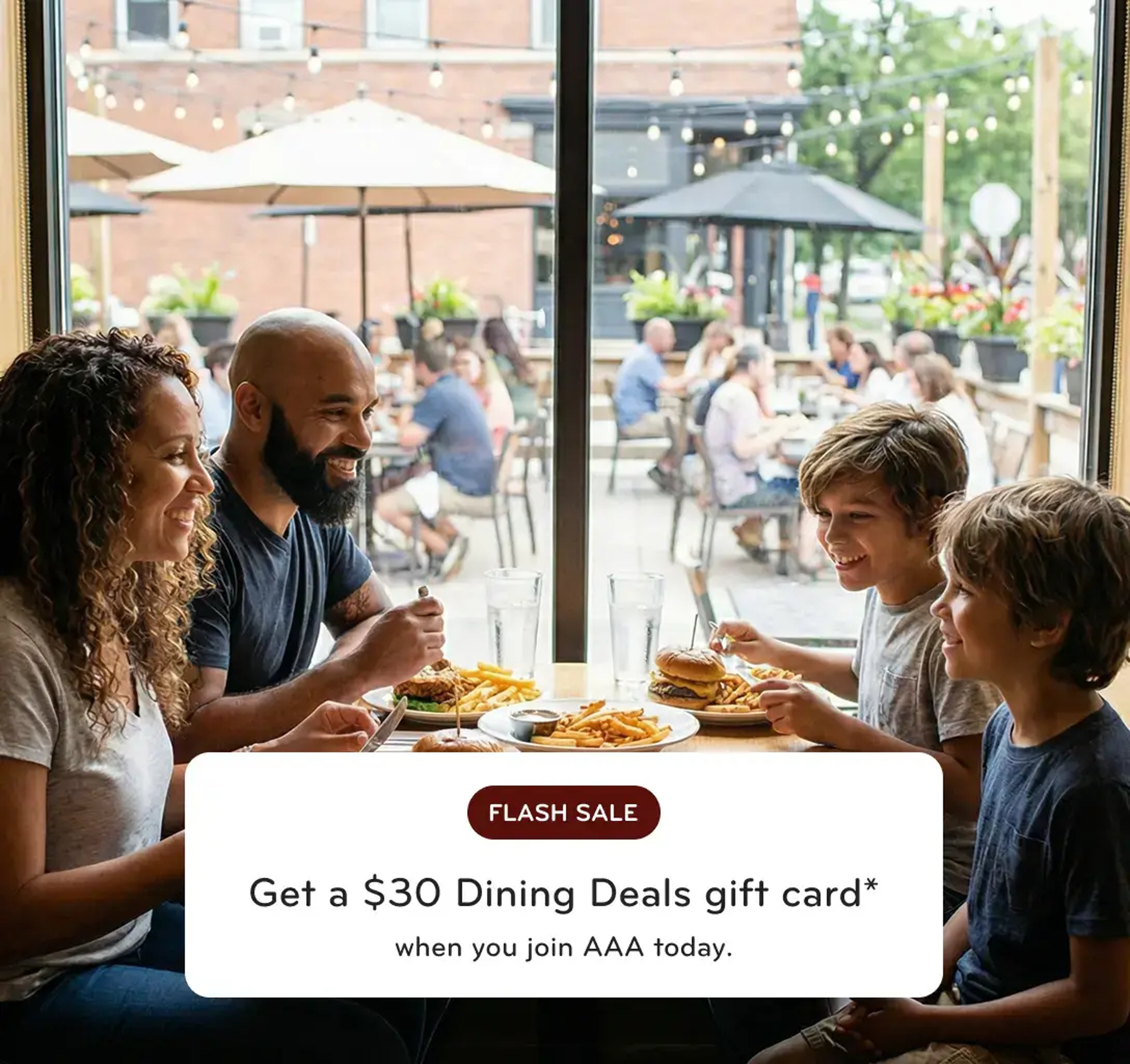 A happy family of four enjoying burgers and pasta at a restaurant table next to a large window overlooking an outdoor patio.