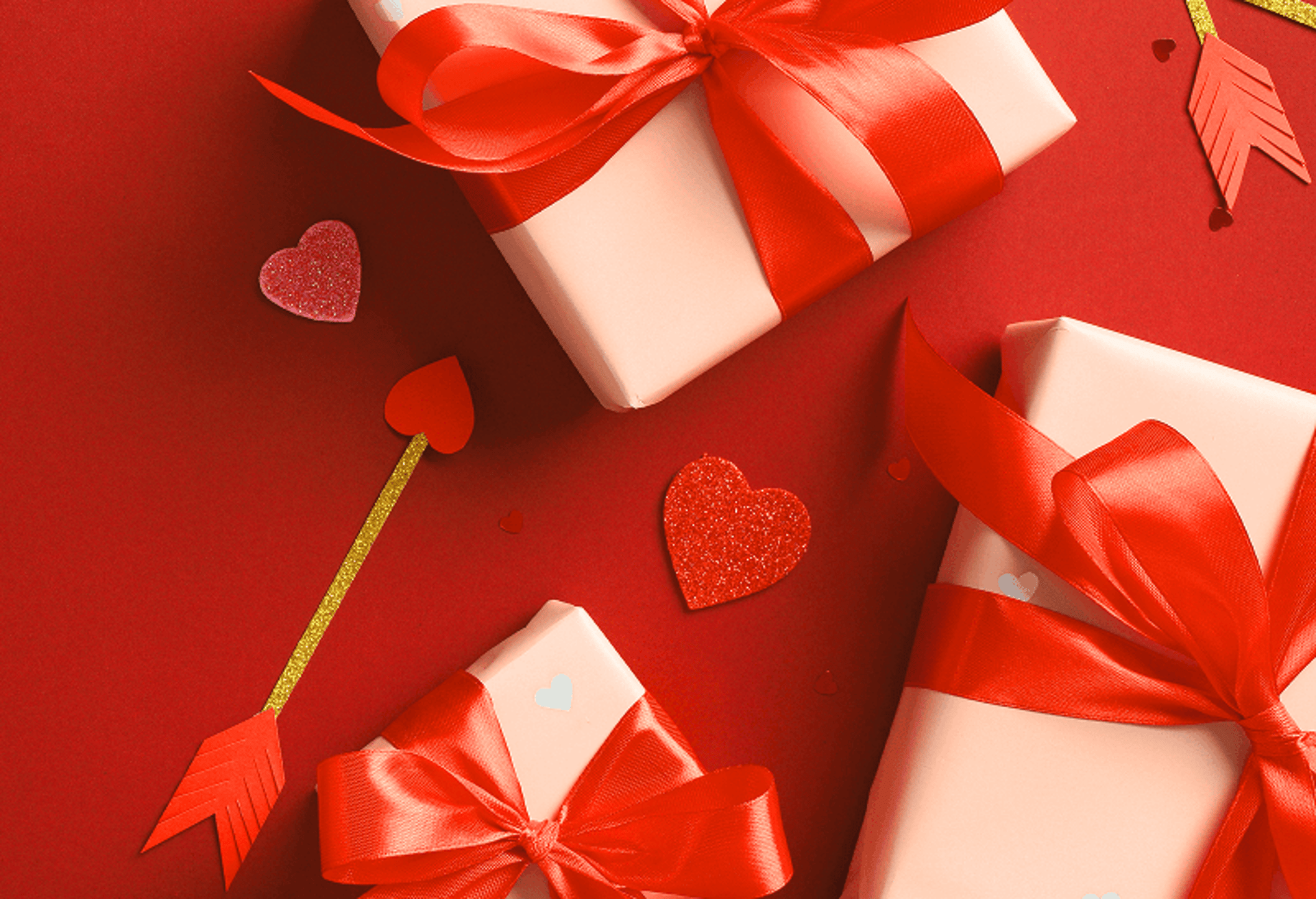 High-angle shot of three rectangular gifts wrapped in pale pink paper and secured with vibrant red satin ribbons. They are arranged on a solid red background. Decorative elements including a gold-glittered Cupid's arrow with a red heart tip and several small red glittery hearts are scattered around the boxes.