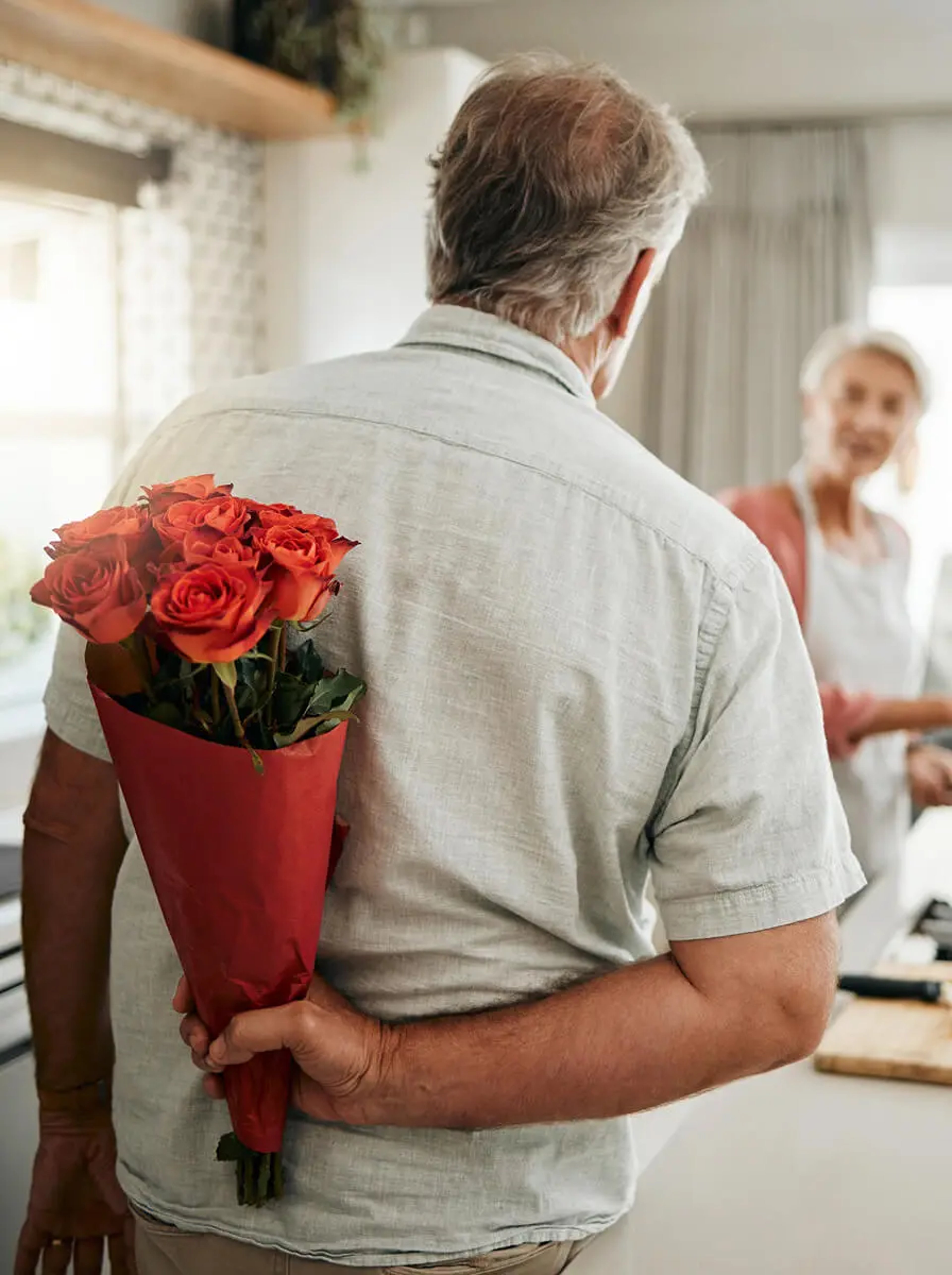 A medium shot from behind an older man with grey hair, wearing a light-colored button-down shirt. He is holding a bouquet of vibrant red roses wrapped in red paper behind his back. In the soft-focus background, an older woman in an apron looks toward him with a smile while standing in a bright, modern kitchen.
