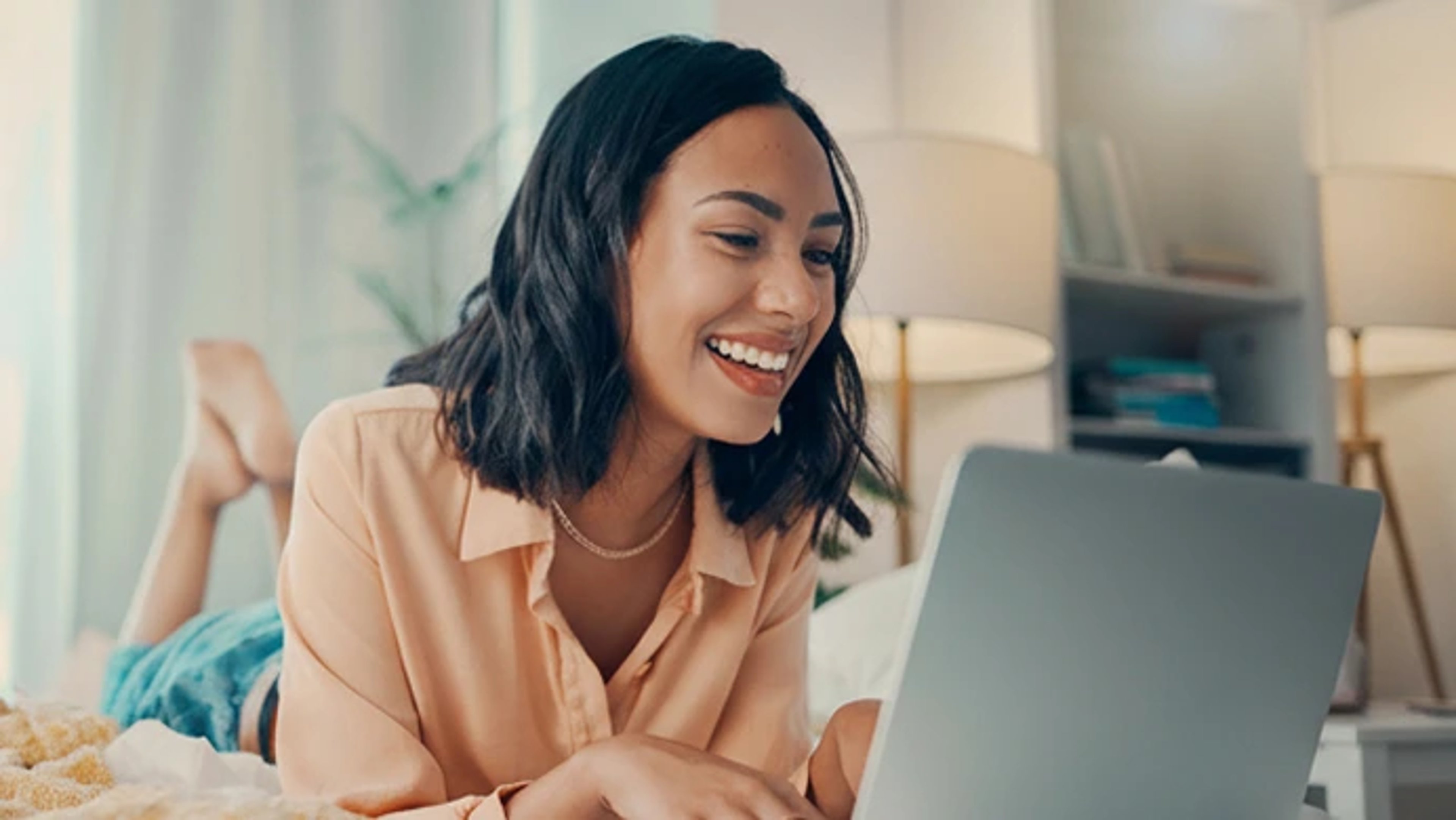 A smiling young woman with dark hair lying on her stomach on a bed while using a laptop in a brightly lit, modern bedroom