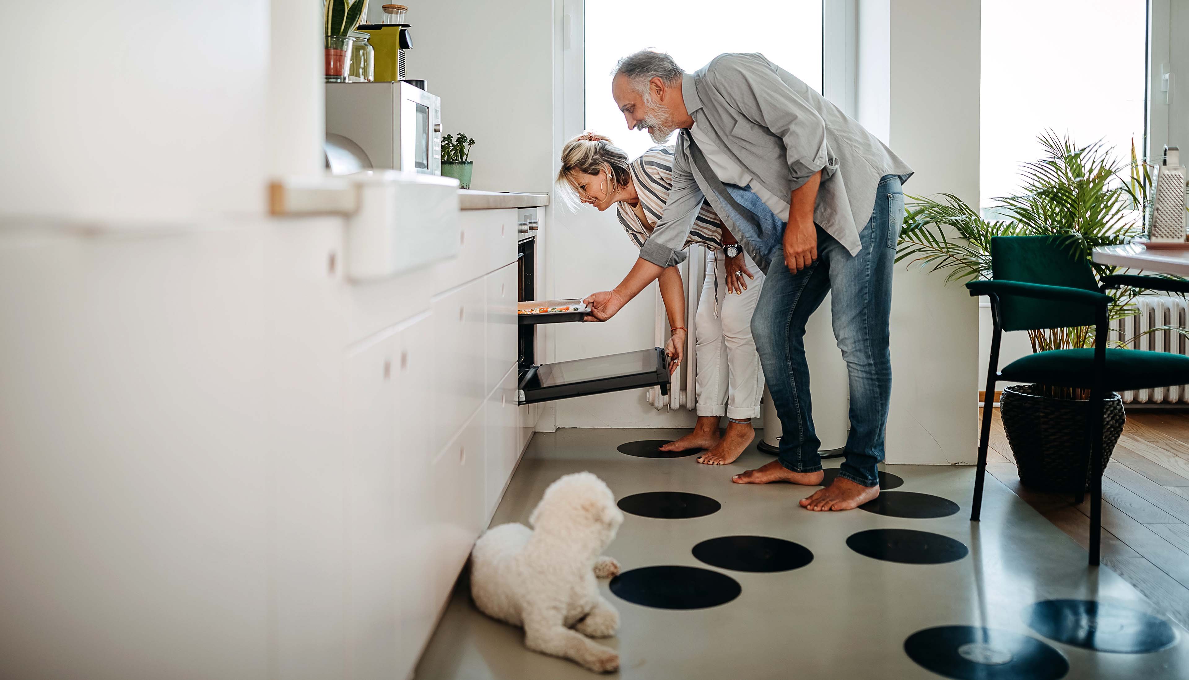 A couple checking an oven in a modern kitchen with a small white dog on the floor.