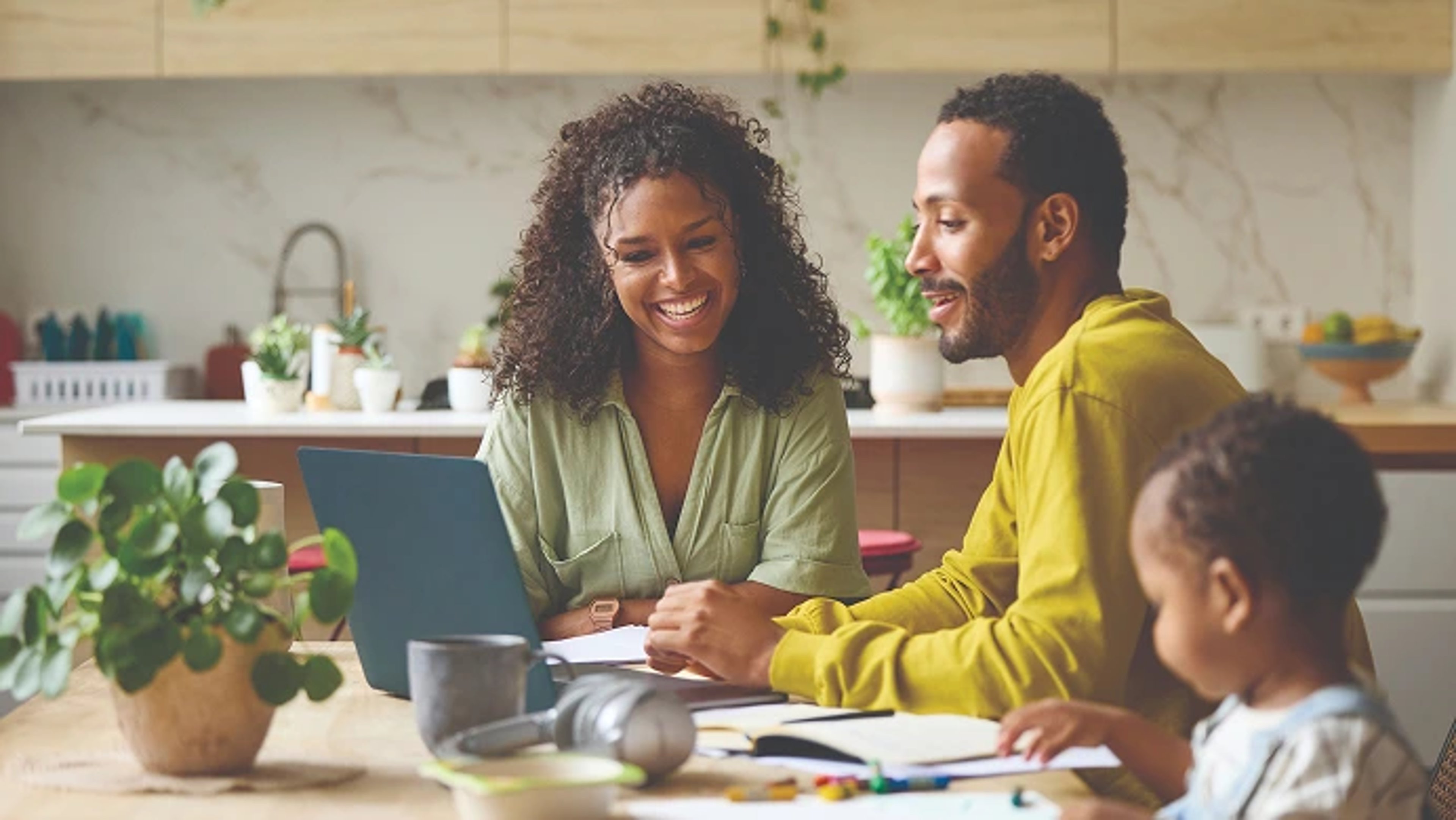 smiling man and woman sitting at a kitchen table looking at a laptop together, with a small child blurred in the foreground and a bright, modern kitchen in the background.