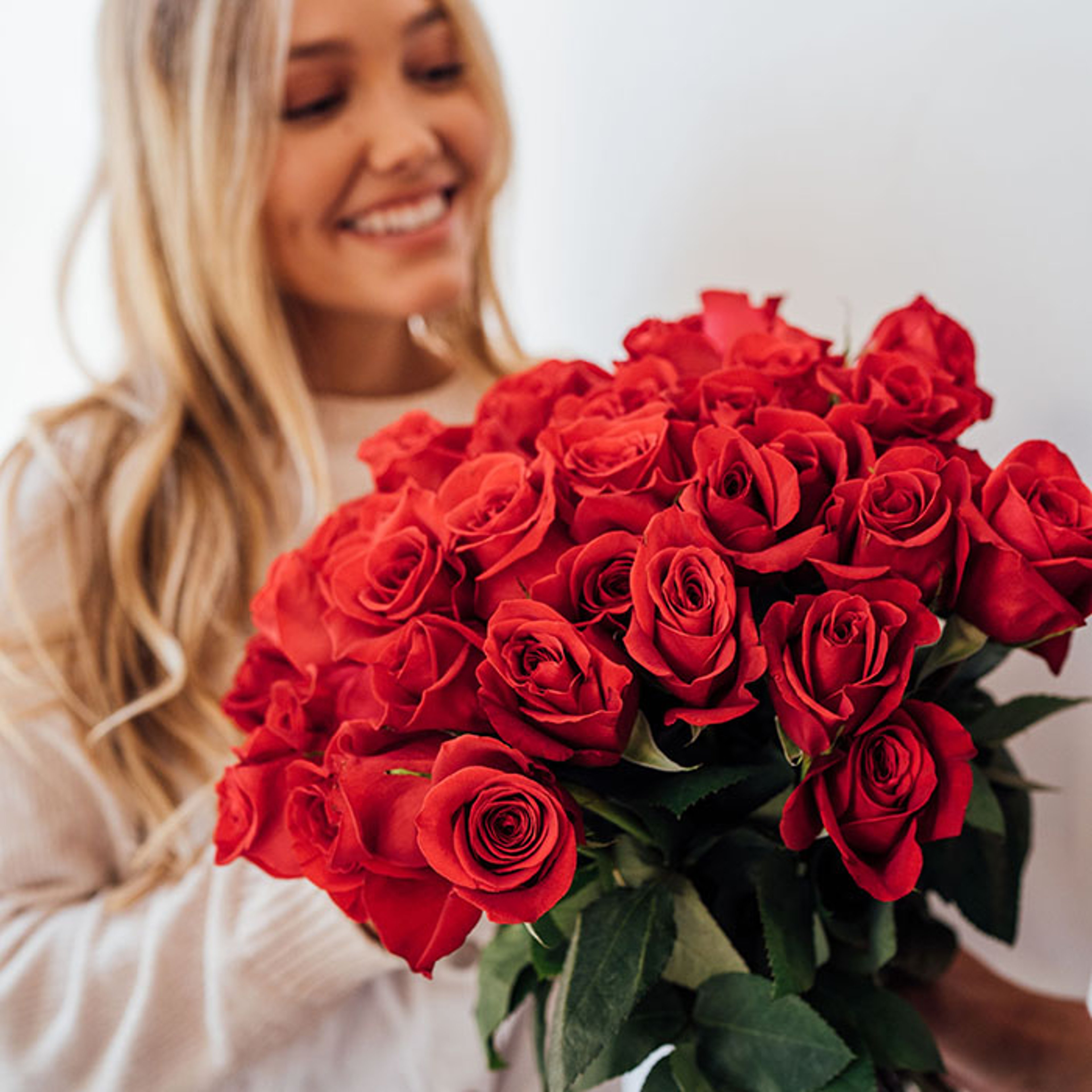 A smiling blonde woman holding a large bouquet of bright red roses in front of her.