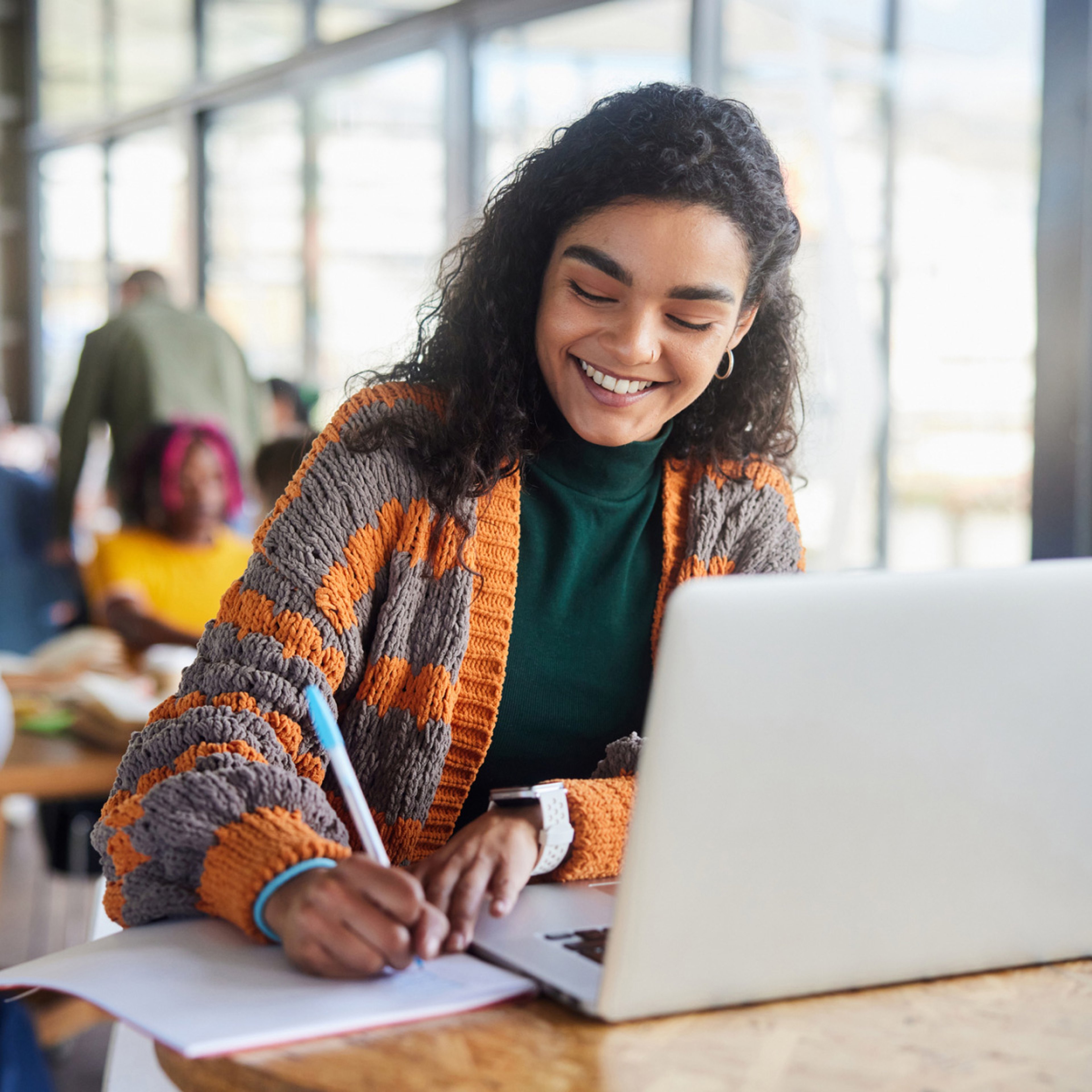 A person smiling while writing in a notebook and using a laptop in a bright room.