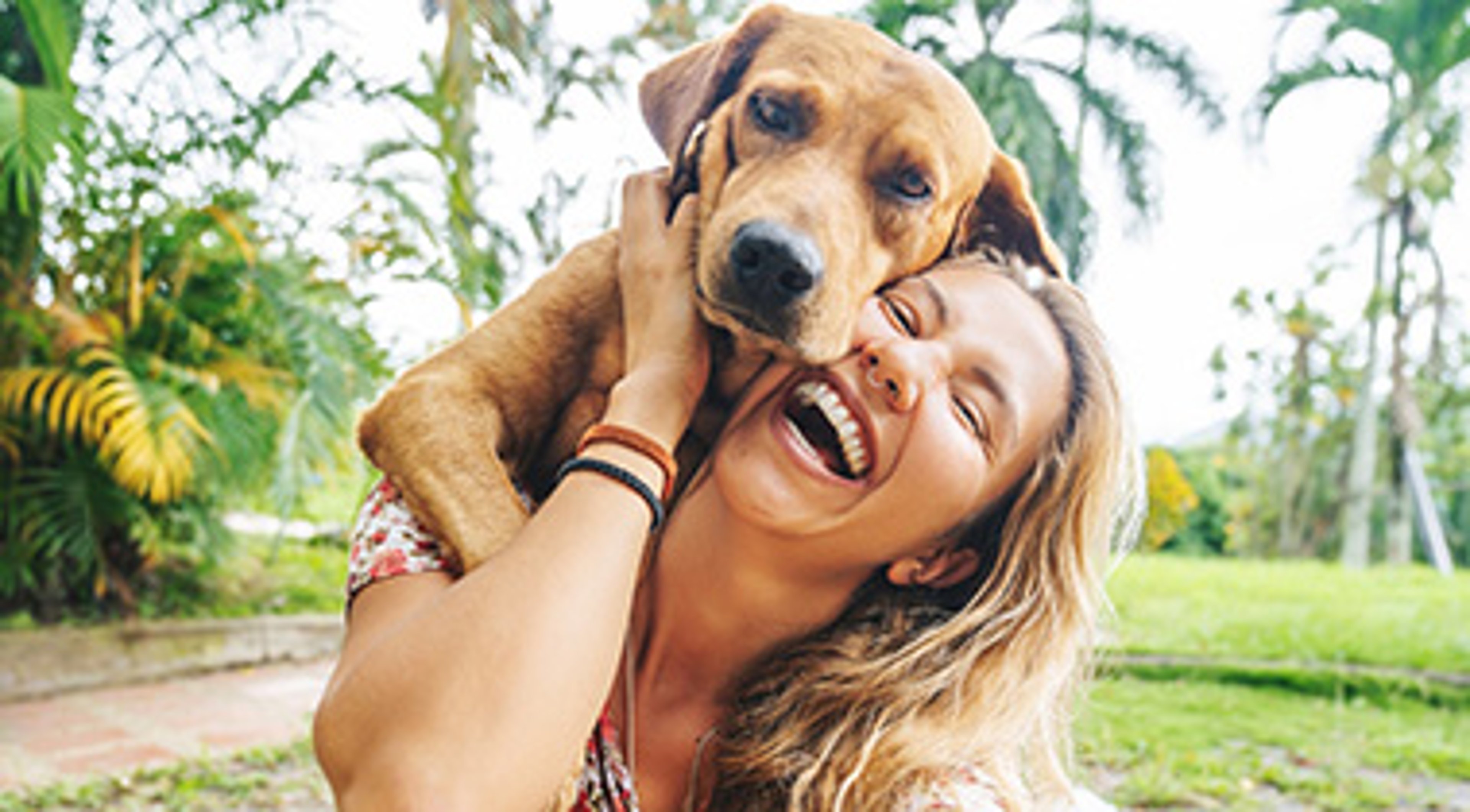 Woman happily holding a dog in her arms, outdoors with trees in the background.