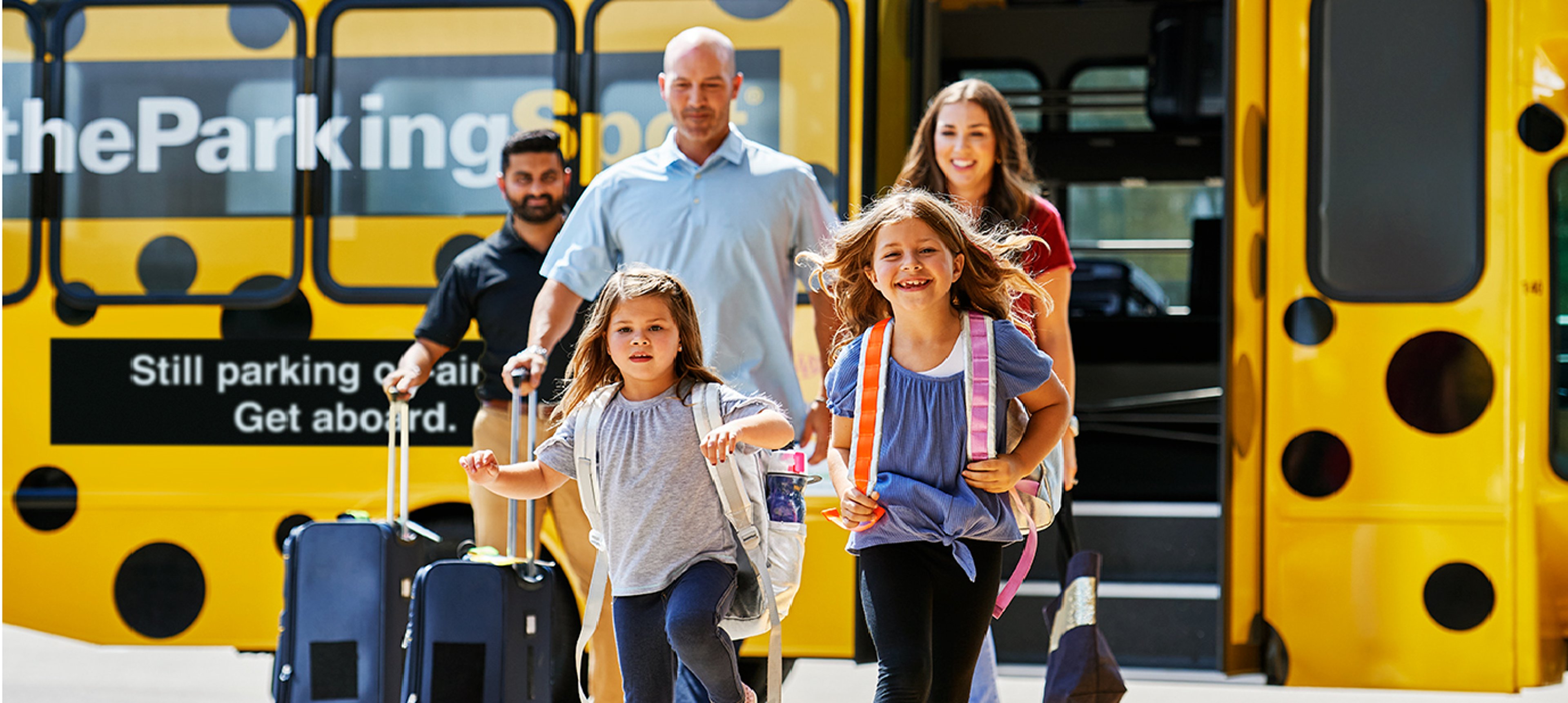 Family of four exiting a yellow bus with black polka dots as The Parking Spot attendant assists with their luggage