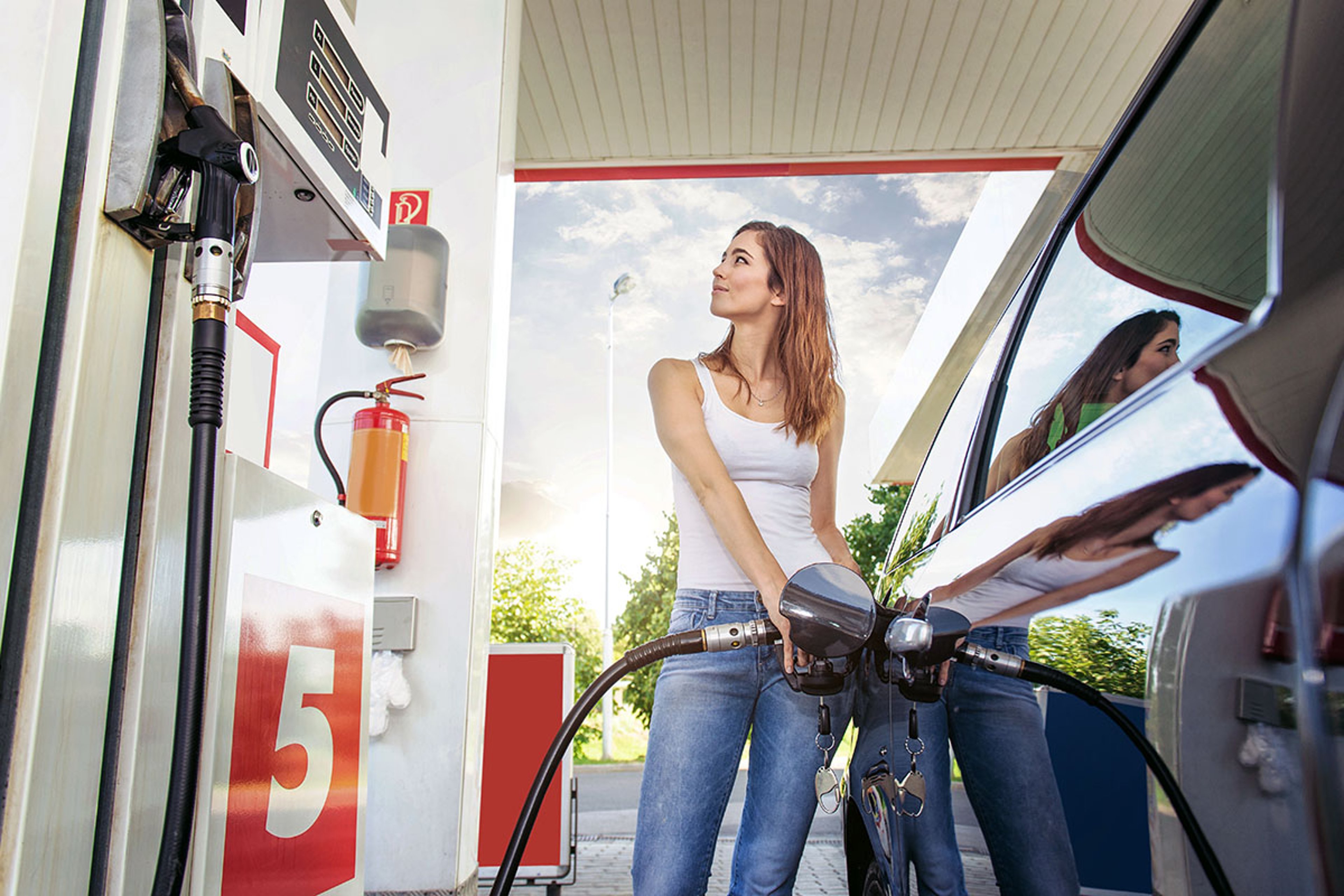 A woman stands at a gas station pump, holding the nozzle as she refuels her car. She is looking upward, and her reflection is visible in the shiny side of the vehicle. The pump is labeled with the number 5, and a fire extinguisher is mounted nearby under the station canopy on a bright day.