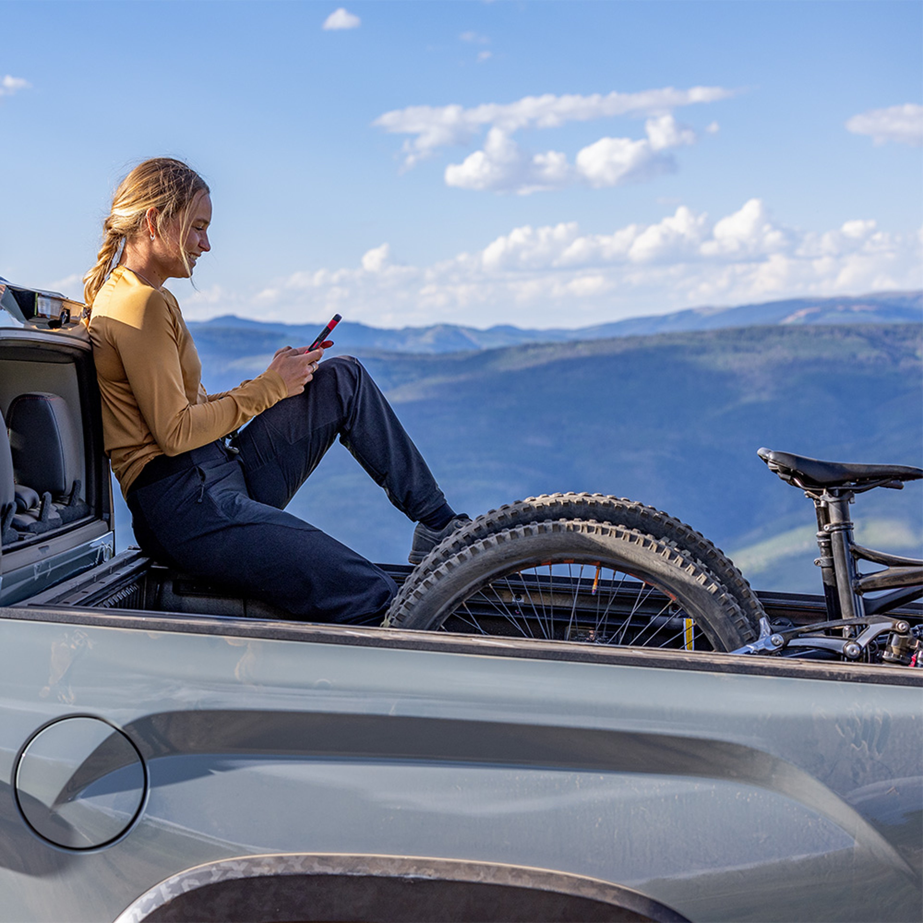 Young woman browsing phone in flatbed of pickup truck with bicycle while on mountain vacation