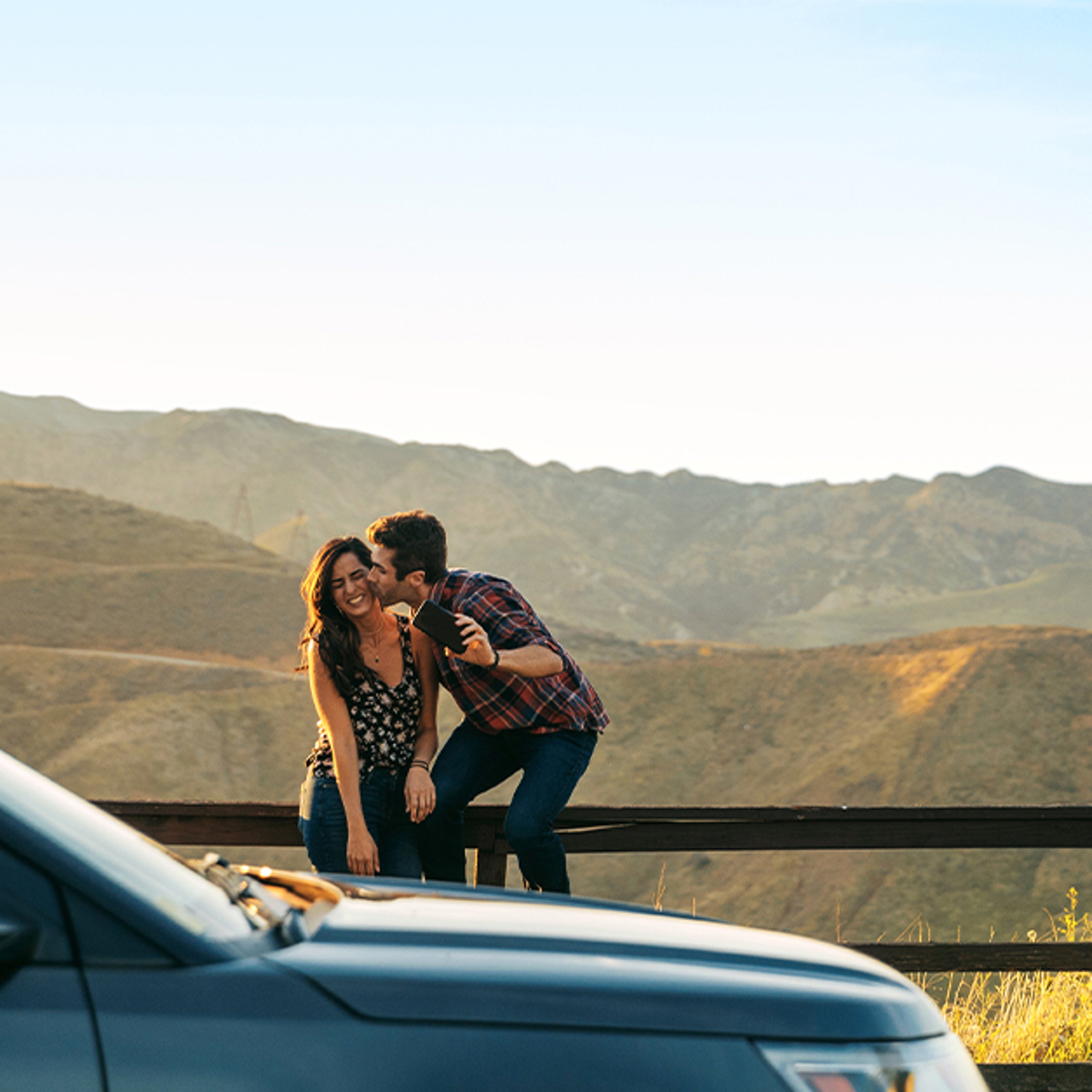 A couple sits on a wooden fence with mountains in the background, while a rental car is parked nearby