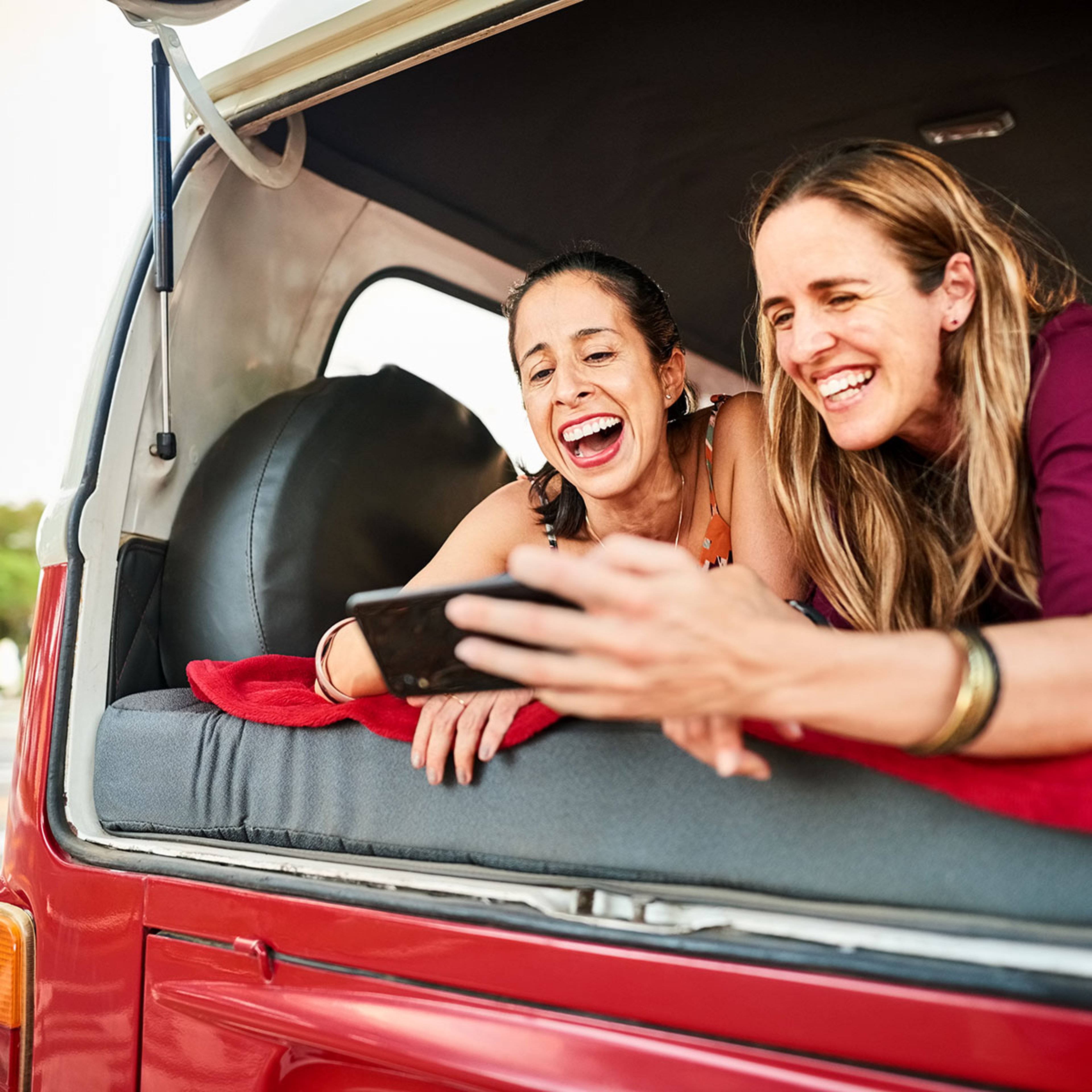 Two young women laughing in trunk of red off-road vehicle as they look at their smartphones