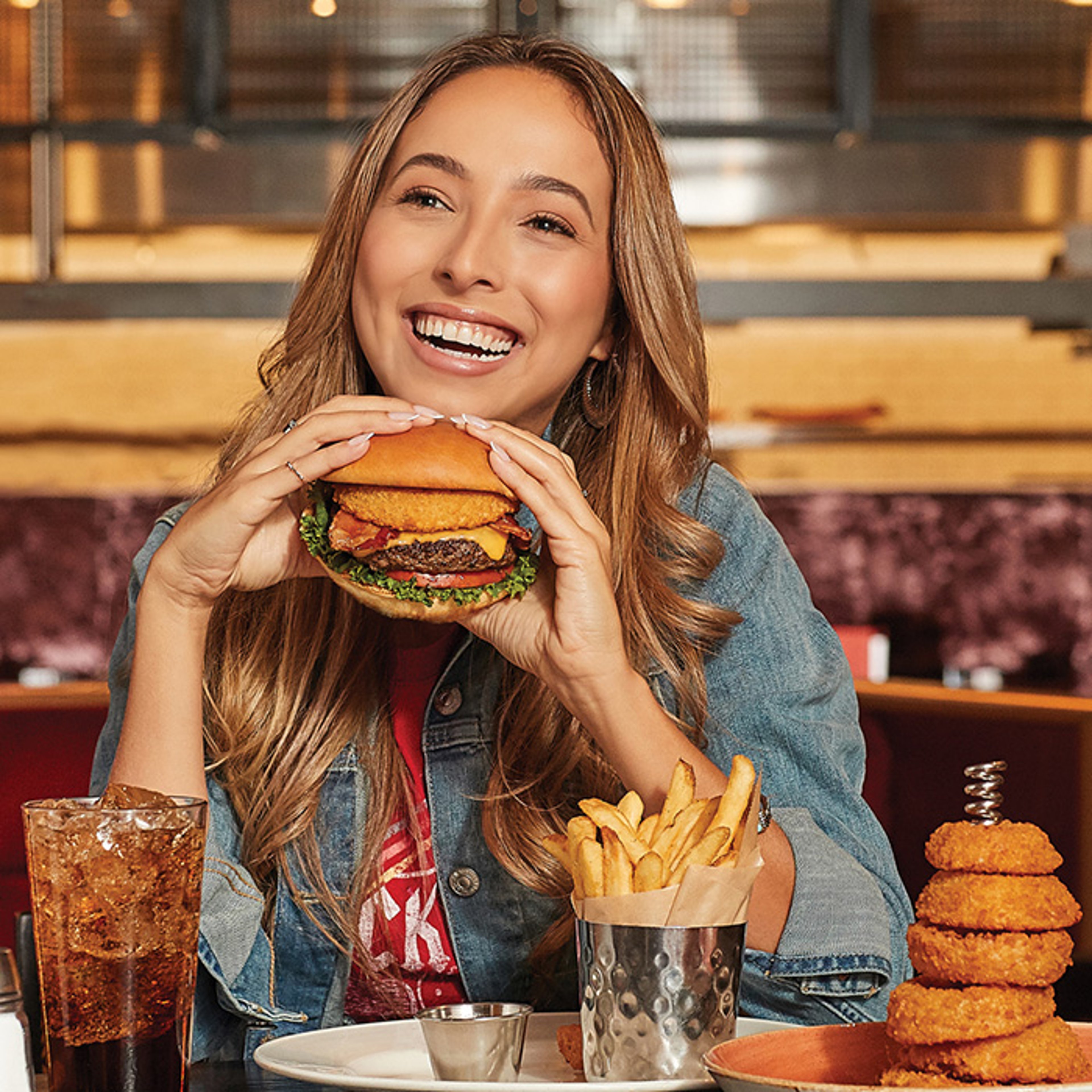 Woman sitting in booth at restaurant smiling as she enjoys eating a burger and fries