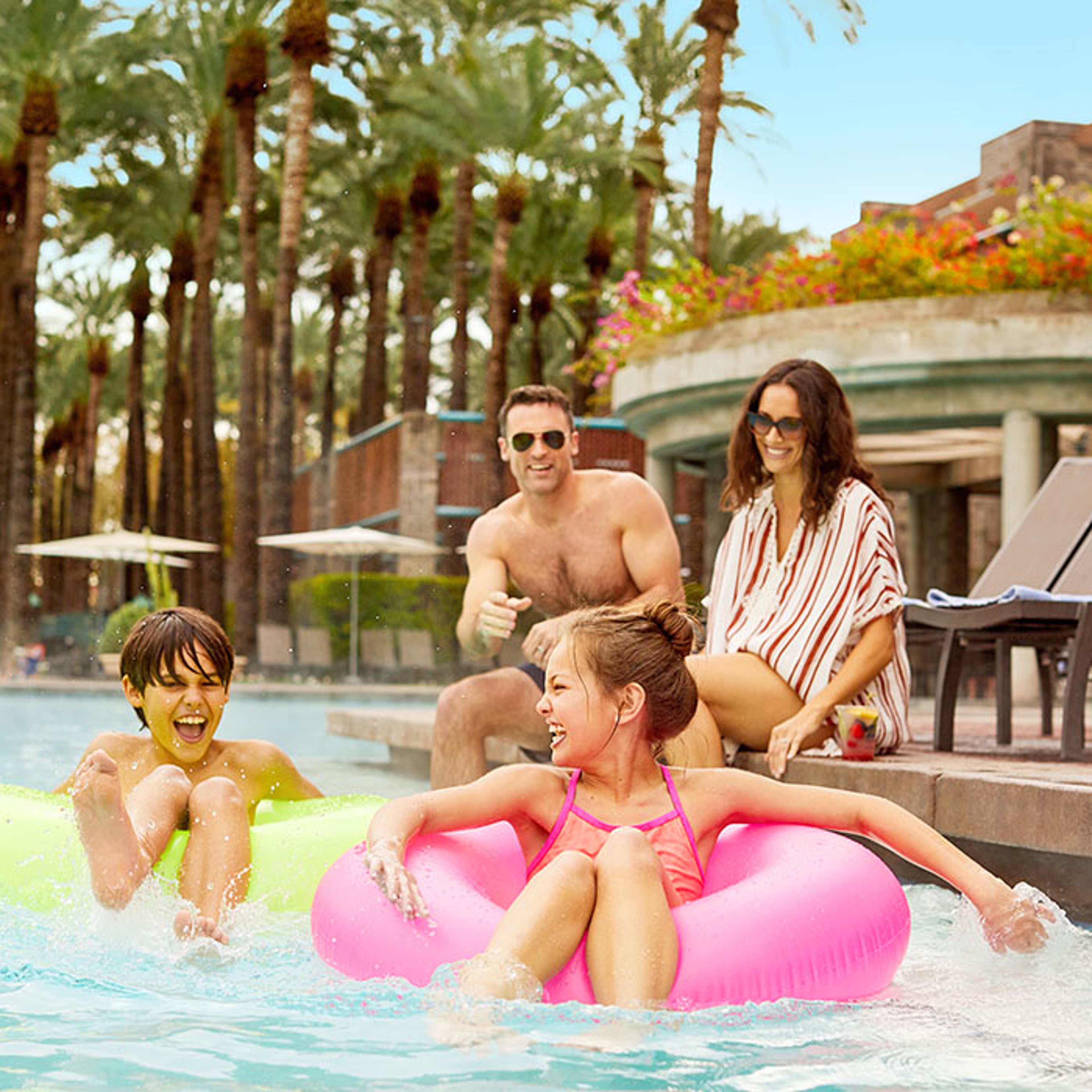 Family laughing with children in tube floats in pool at beach resort with palm trees