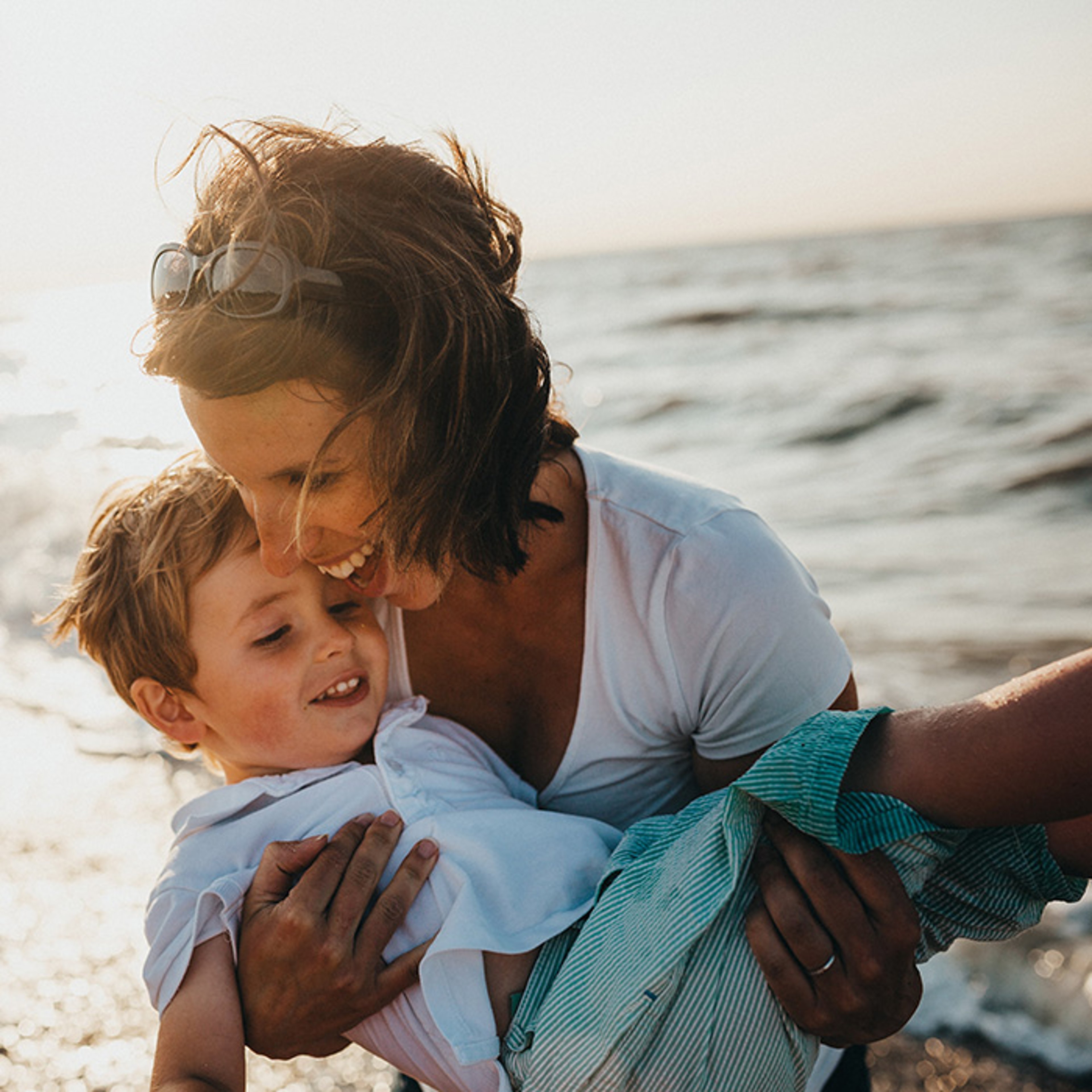 Mother holding son laughing at the beach on sunny day with ocean in background
