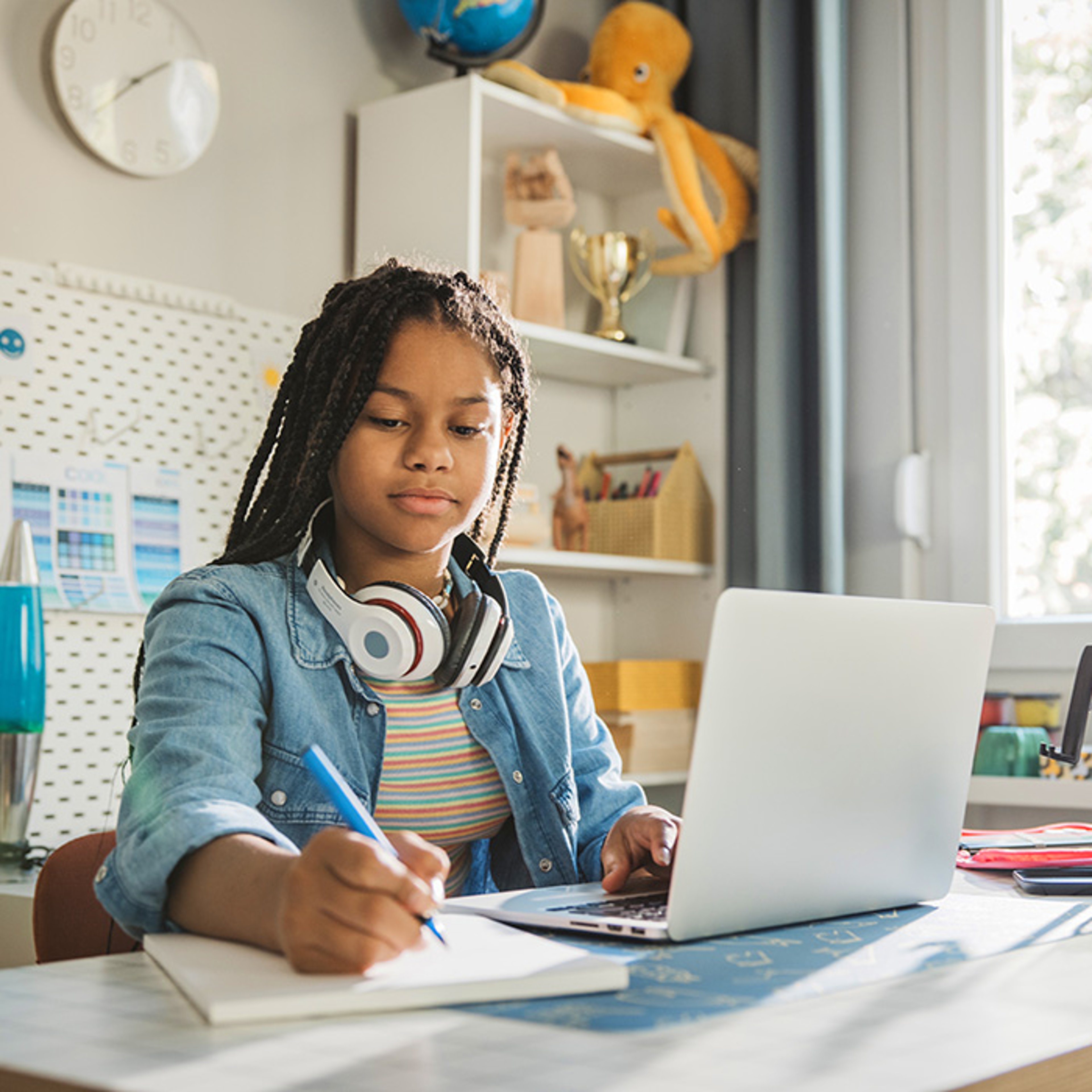 Young girl doing schoolwork on laptop while writing notes in her bedroom