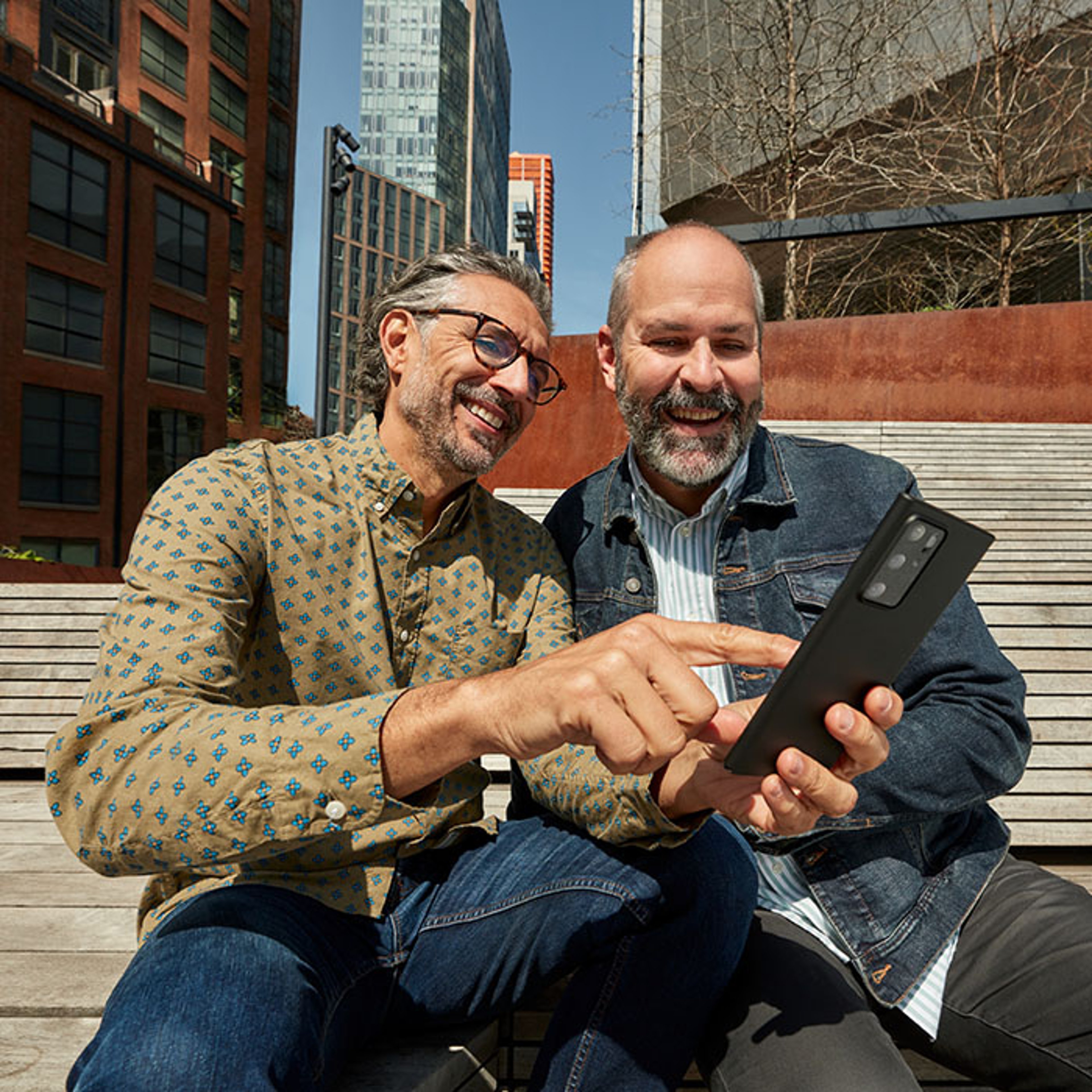 Male couple sitting on stairs of city building happily looking at their mobile phone