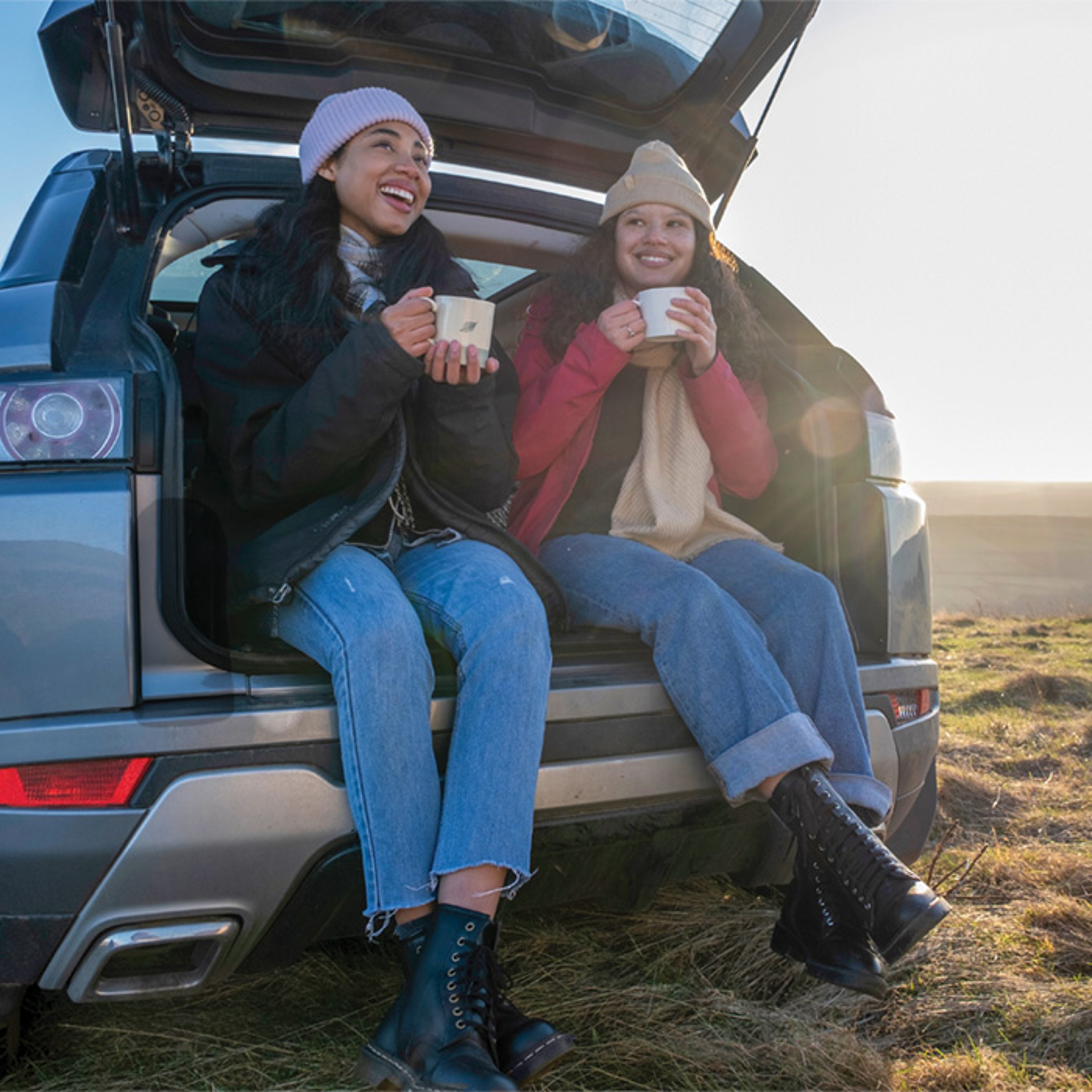 Two smiling young women sipping coffee from mugs while sitting in trunk of rental SUV on fall vacation