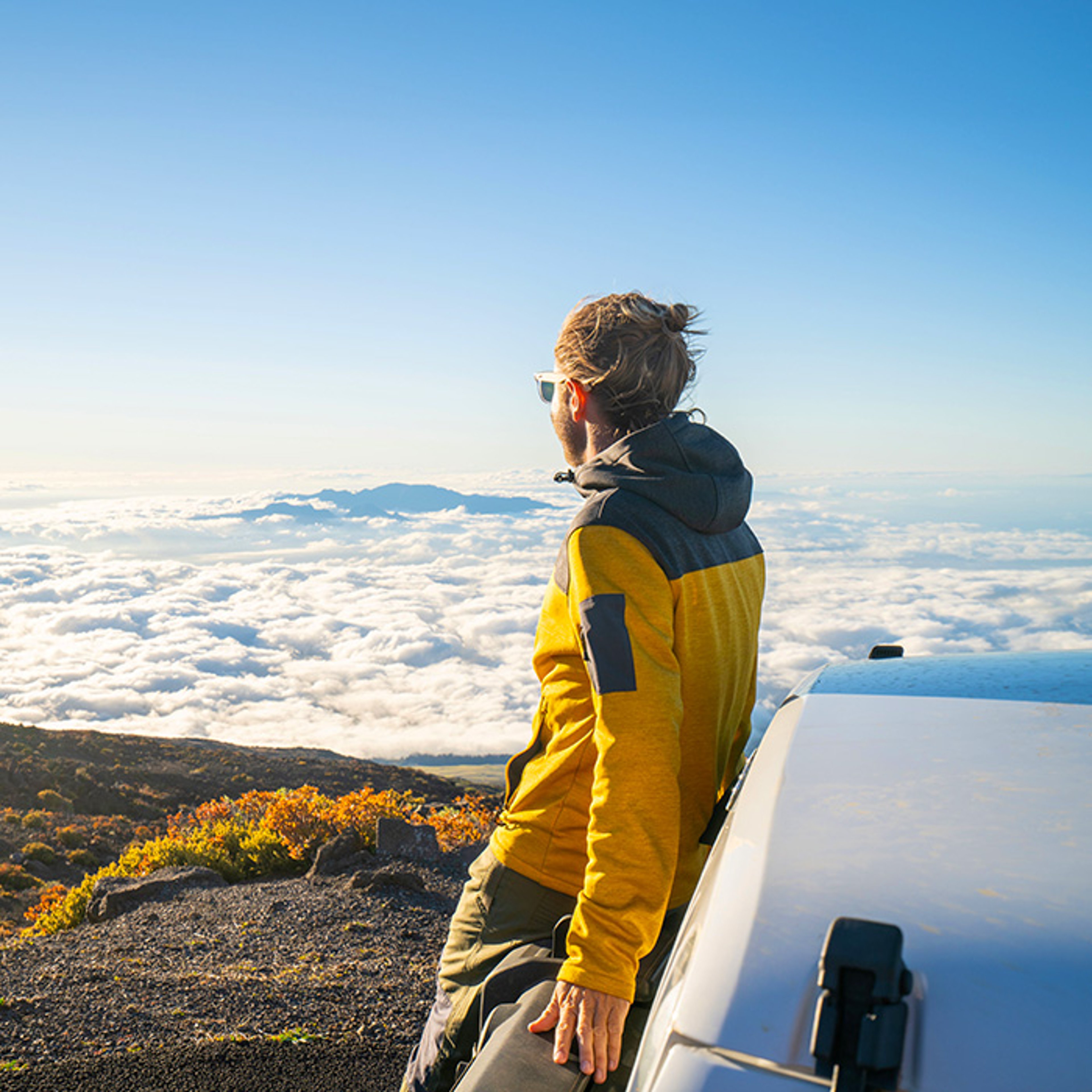 Man standing against rental car atop mountain with view of clouds below on sunny day