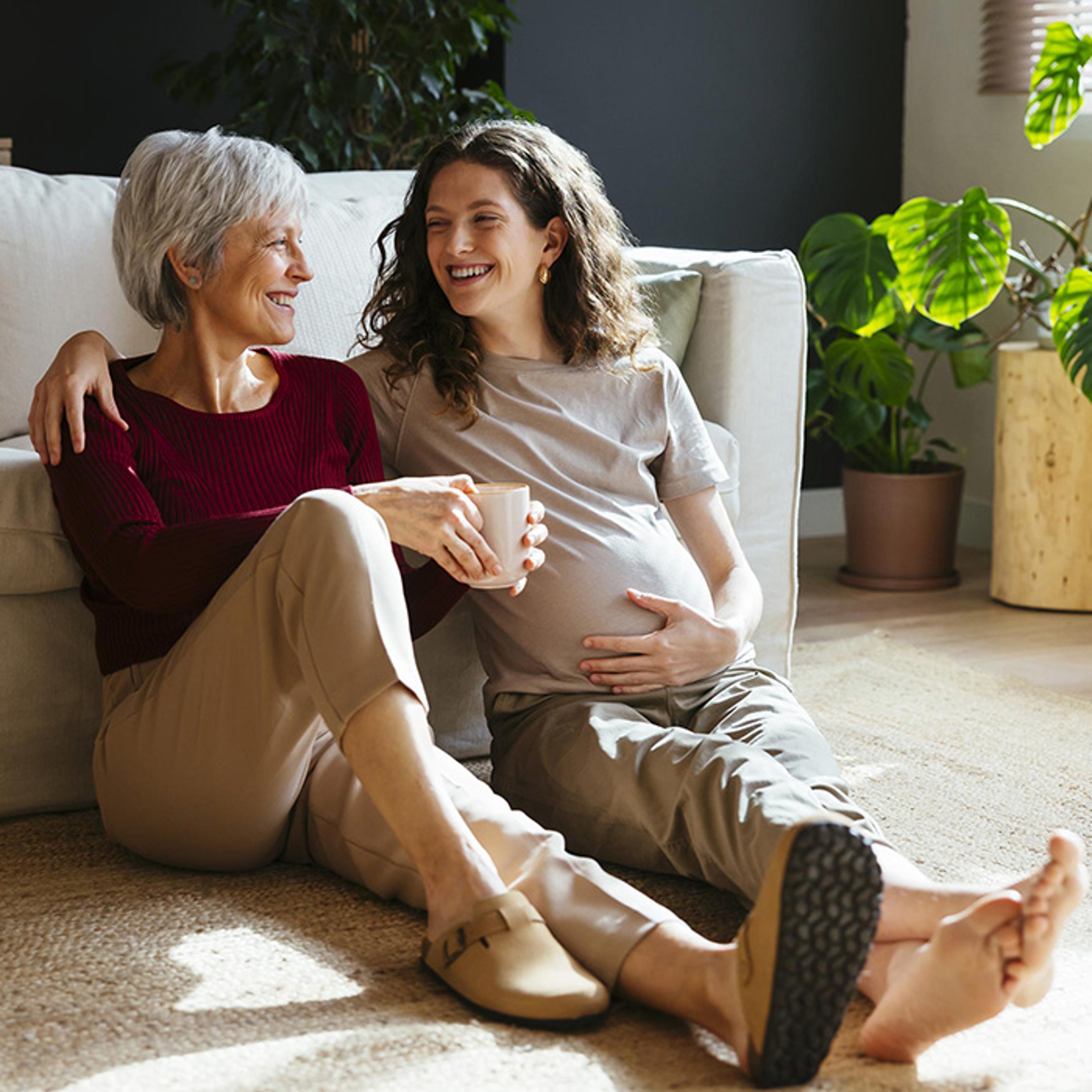 Senior woman and pregnant daughter laughing together as they sit in home with plant in background