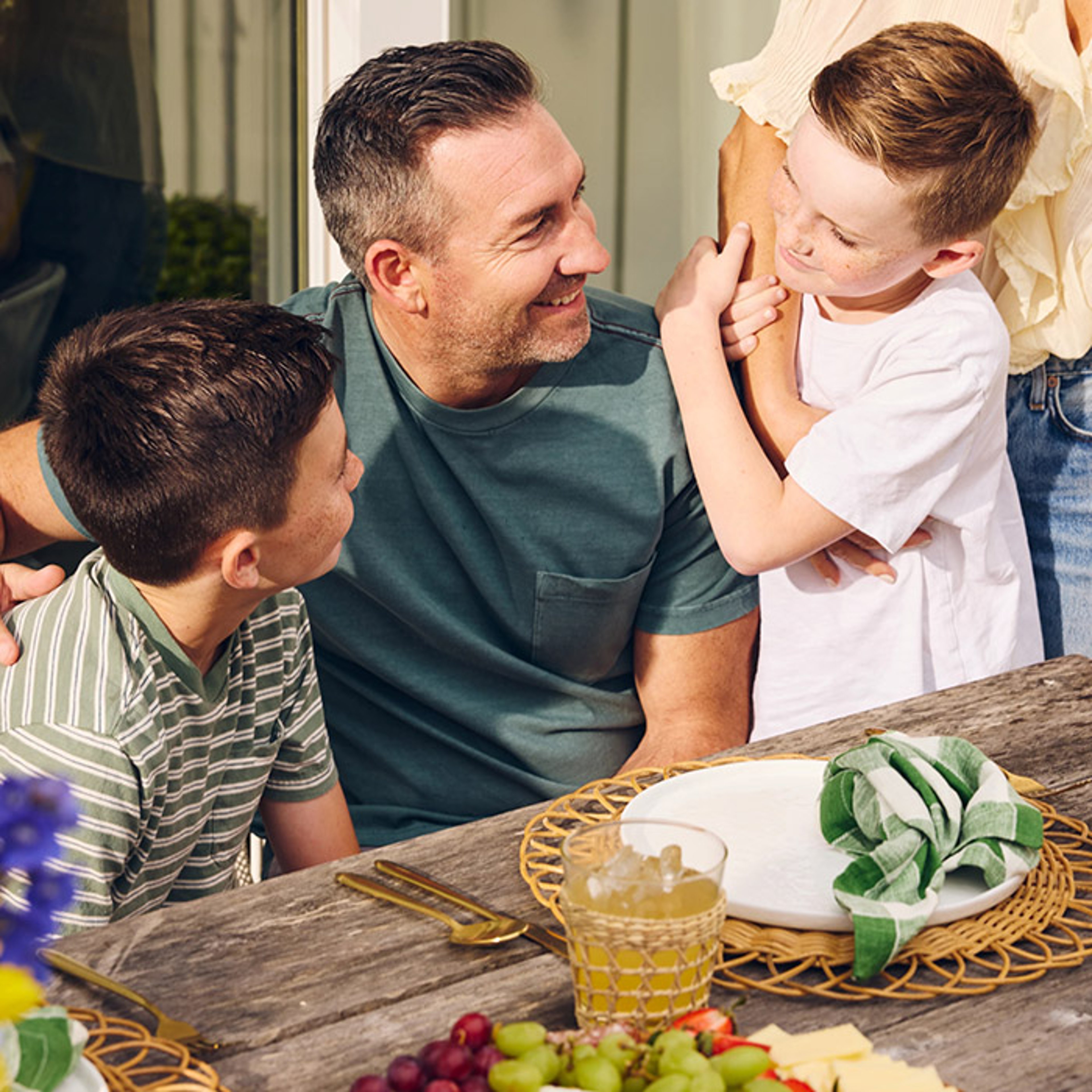 A smiling man sits at a table with two boys, surrounded by a display of fruits and flowers.