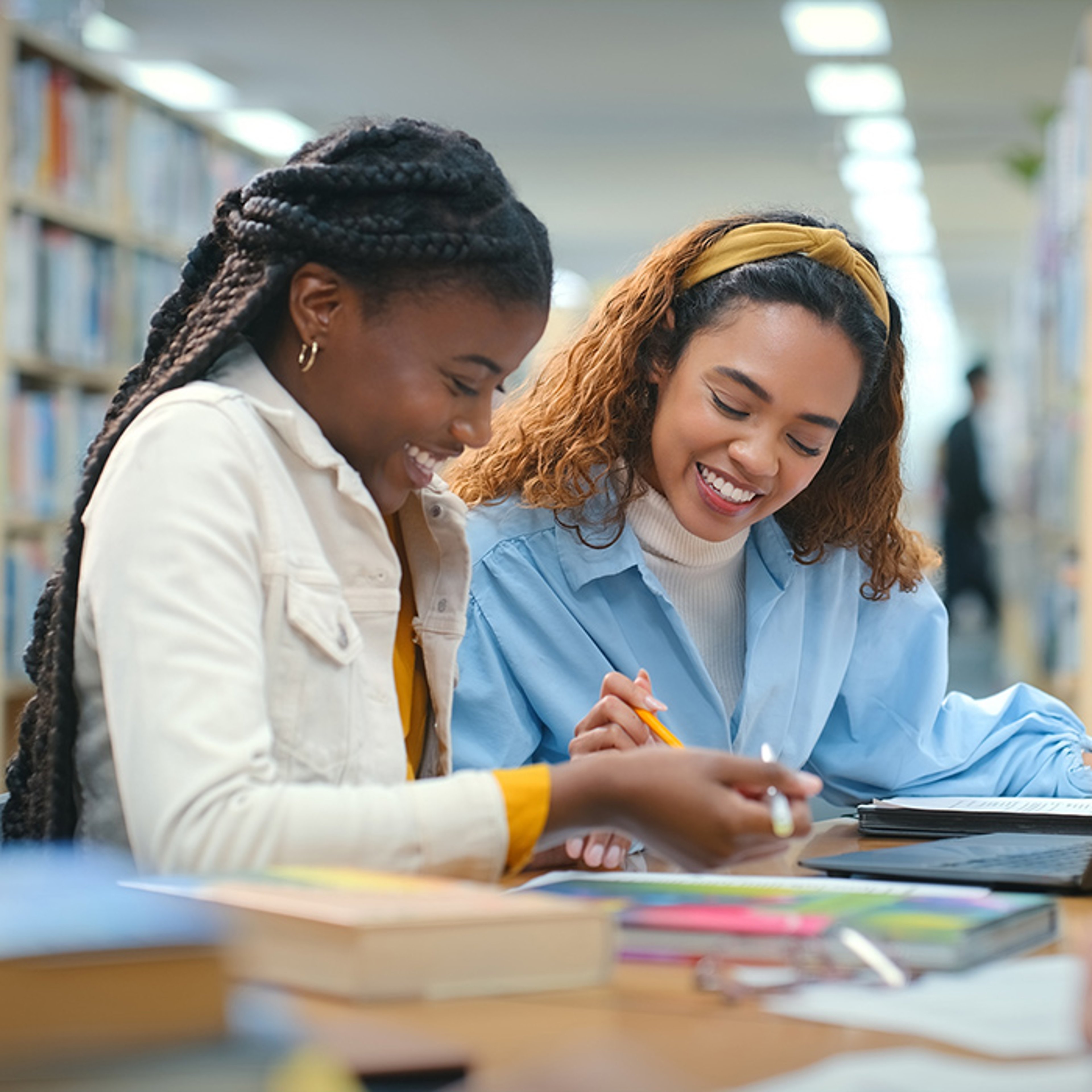 Two women studying together at a library table with books and papers scattered around.