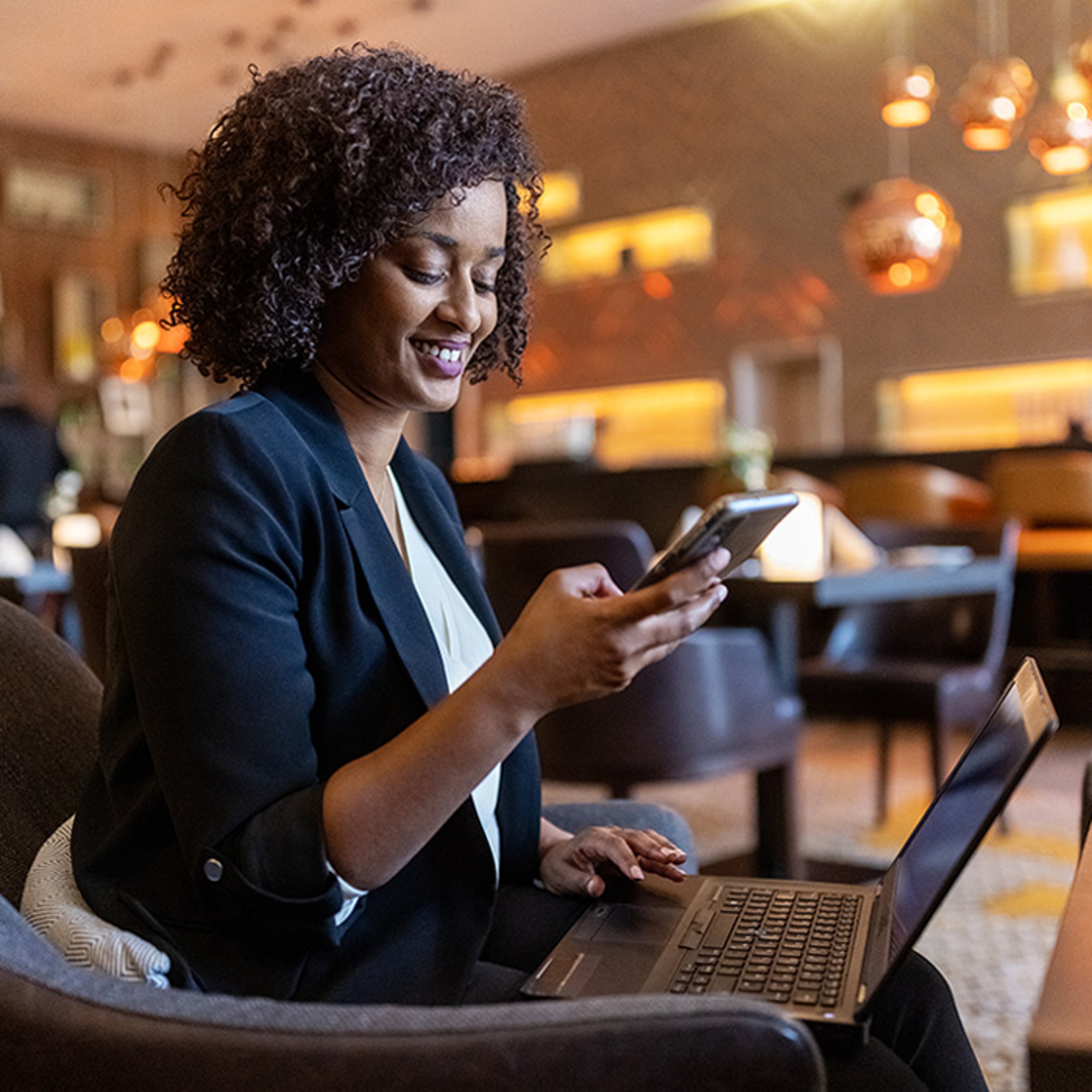 A smiling woman in a black blazer uses a smartphone with a laptop on her lap, in a warmly lit lounge.