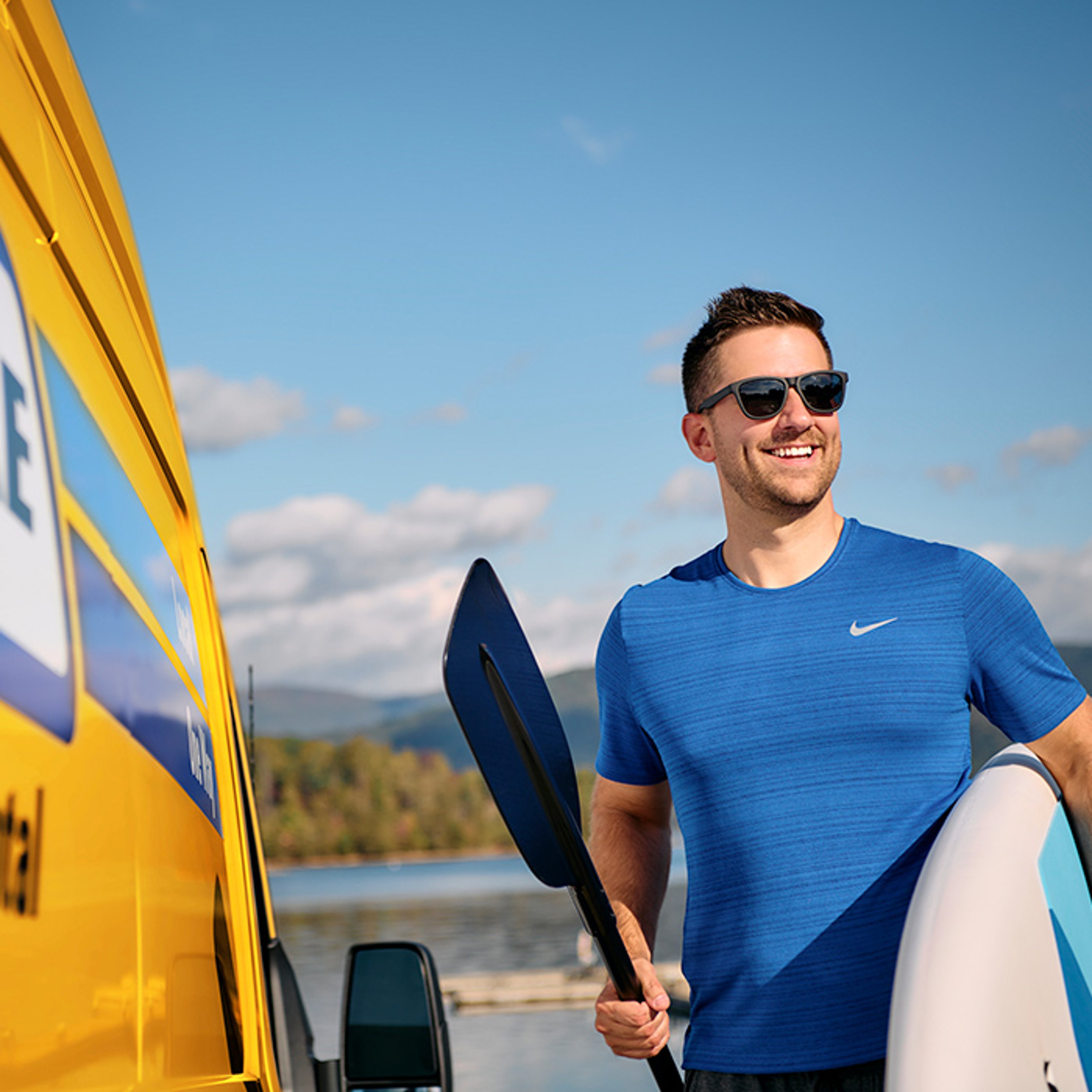 Man in a blue shirt holding a paddle and surfboard next to a yellow vehicle with a scenic lake and mountains in the background.