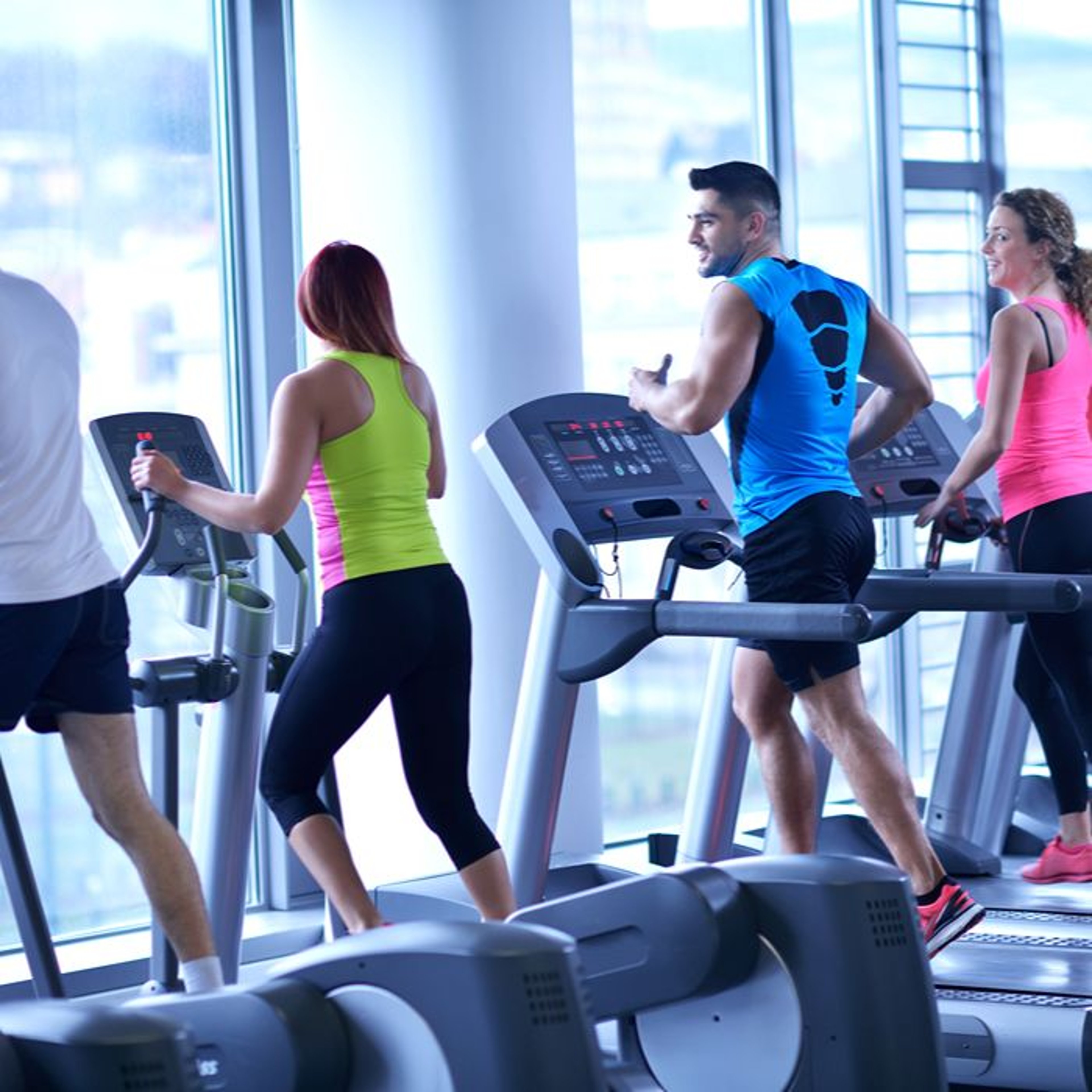 People exercising on treadmills in a bright gym, wearing colorful athletic wear, with large windows in the background.