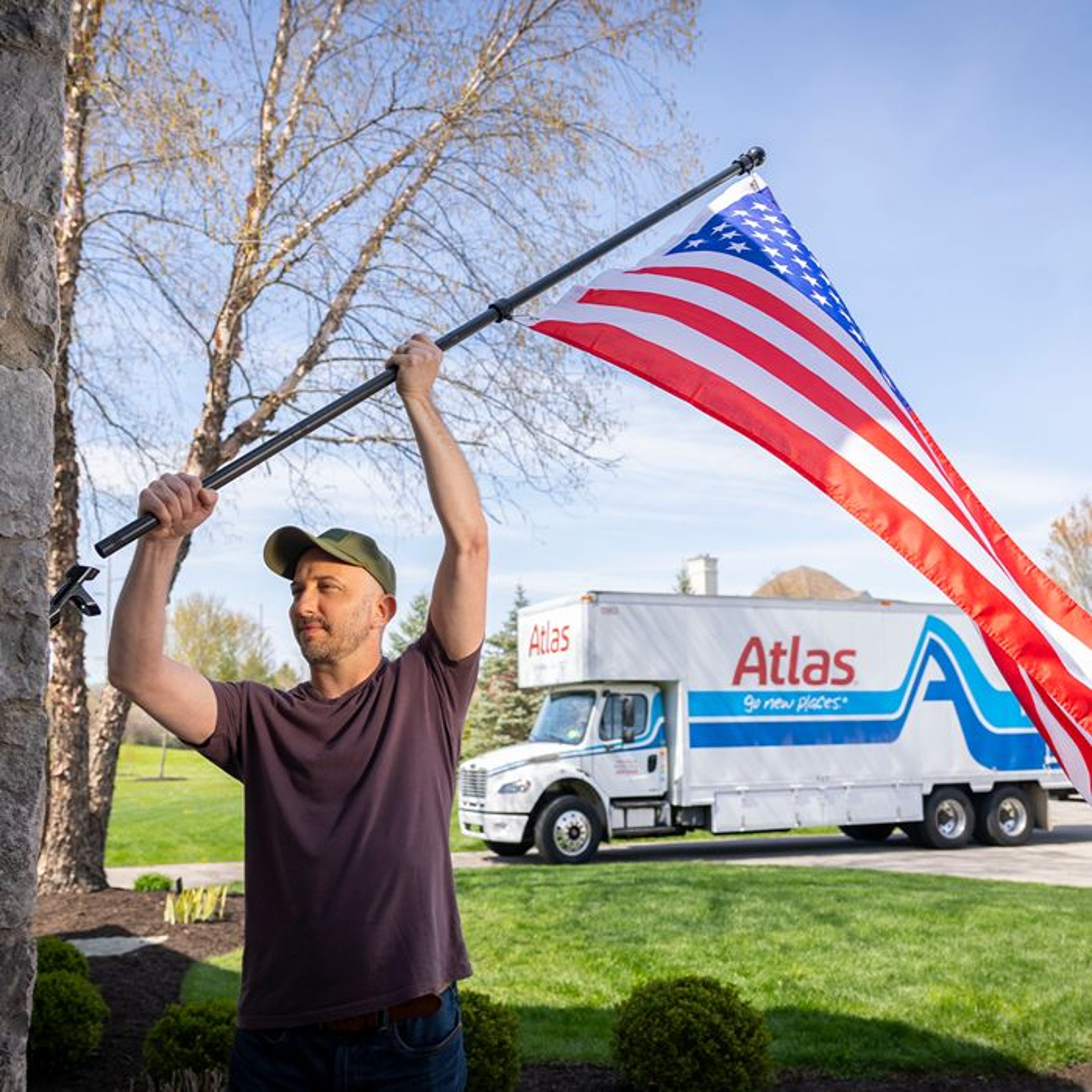 A man raises an American flag outside a building, with an Atlas moving truck parked in the background.