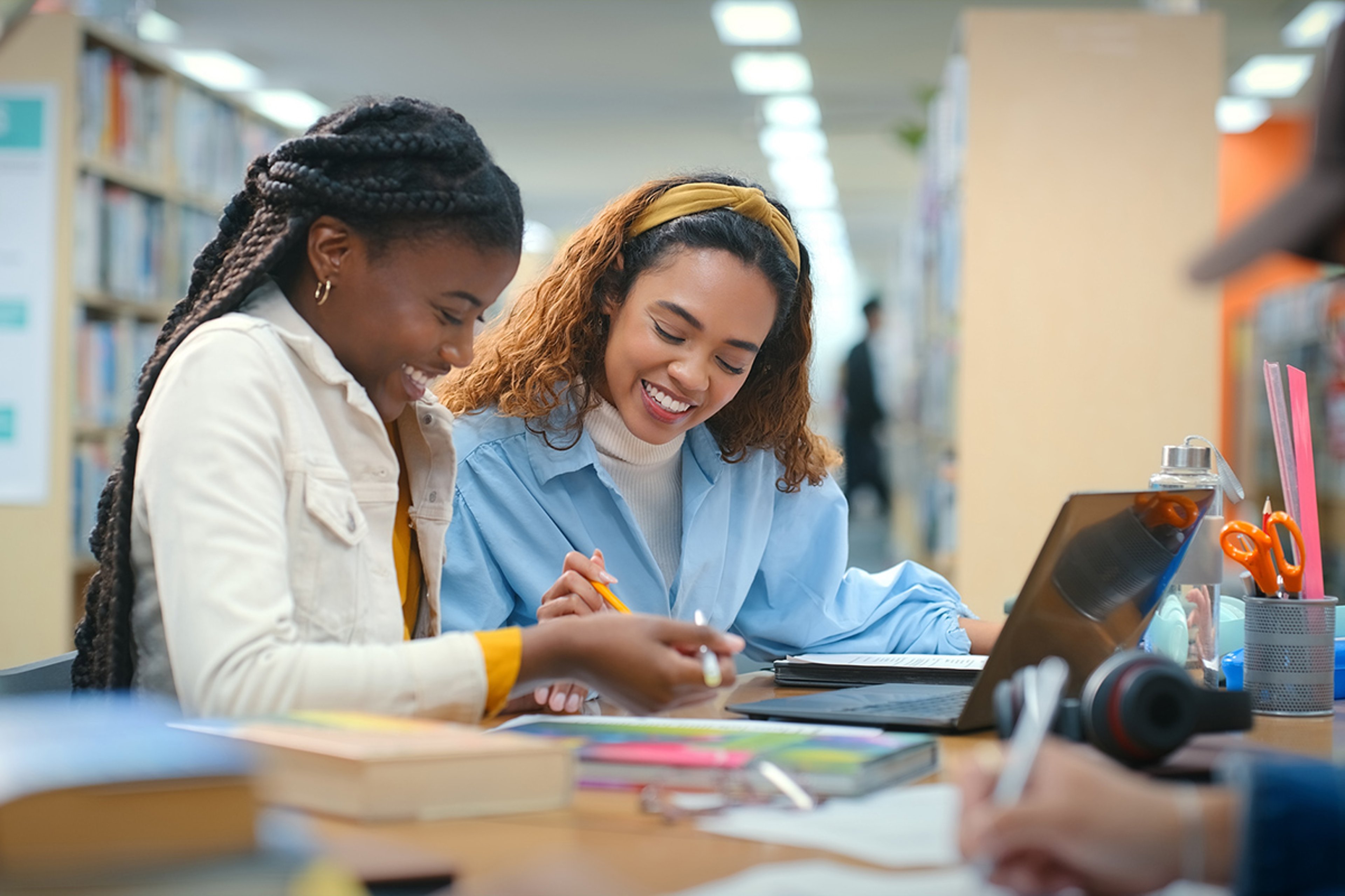 Two young women studying together in a library, smiling and engaging over notebooks and a laptop on their table.