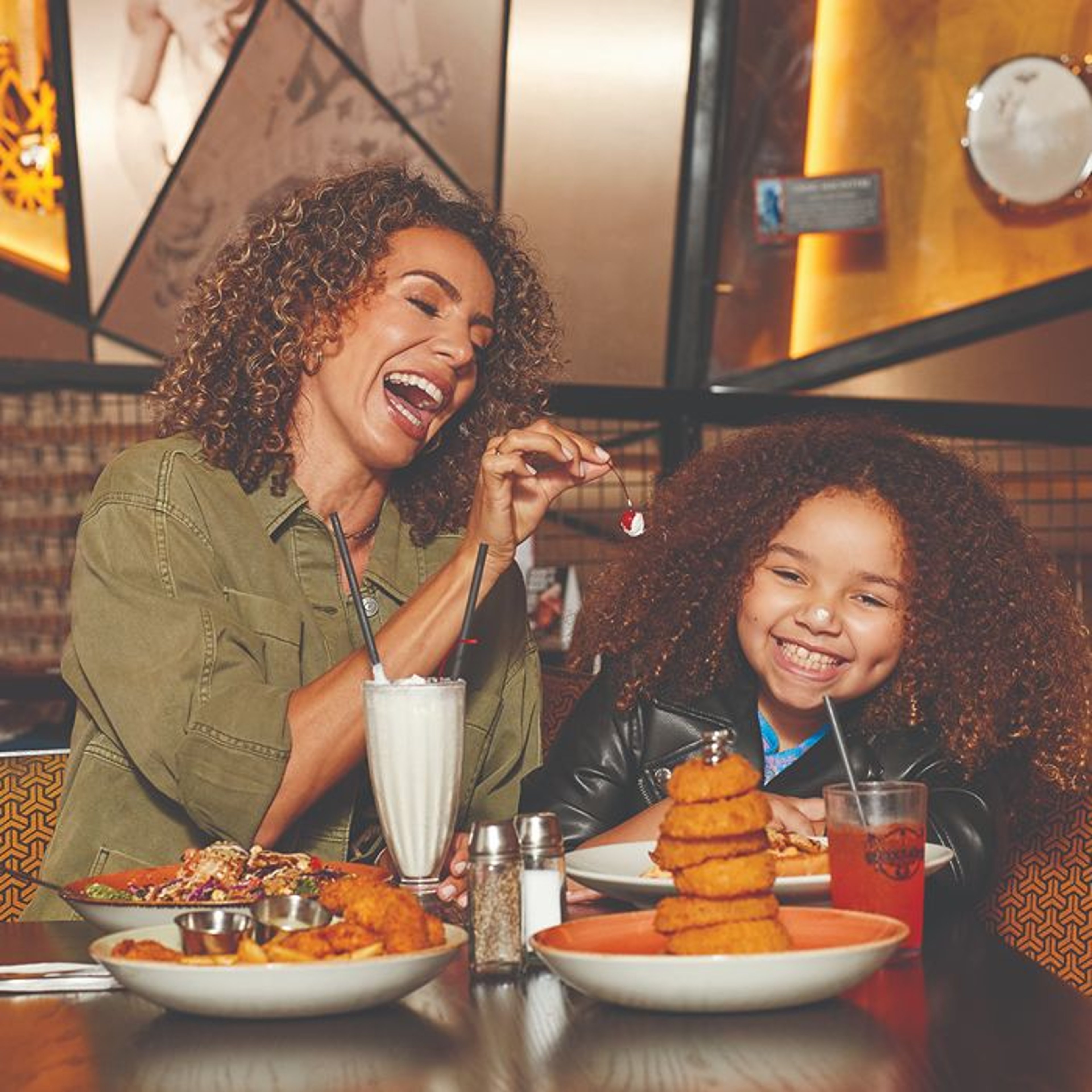 A mother and daughter laugh together at a restaurant, as the mother playfully offers a cherry to the daughter. Multiple plates of food, including a milkshake and fried items, are visible on the table.