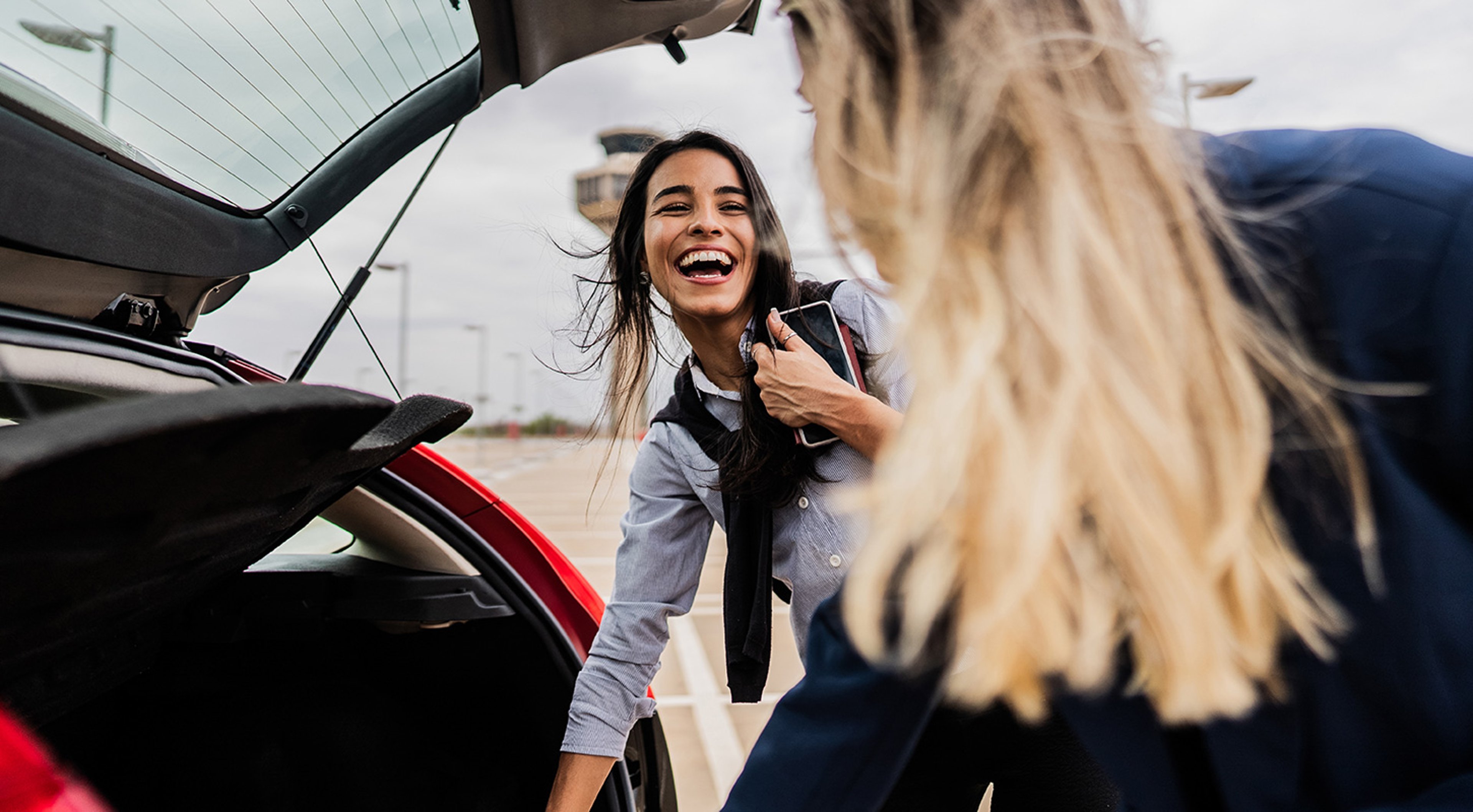 Two women in a parking lot, one smiling and holding a smartphone, while the other retrieves items from the trunk of a red car. An airport control tower is visible in the background.