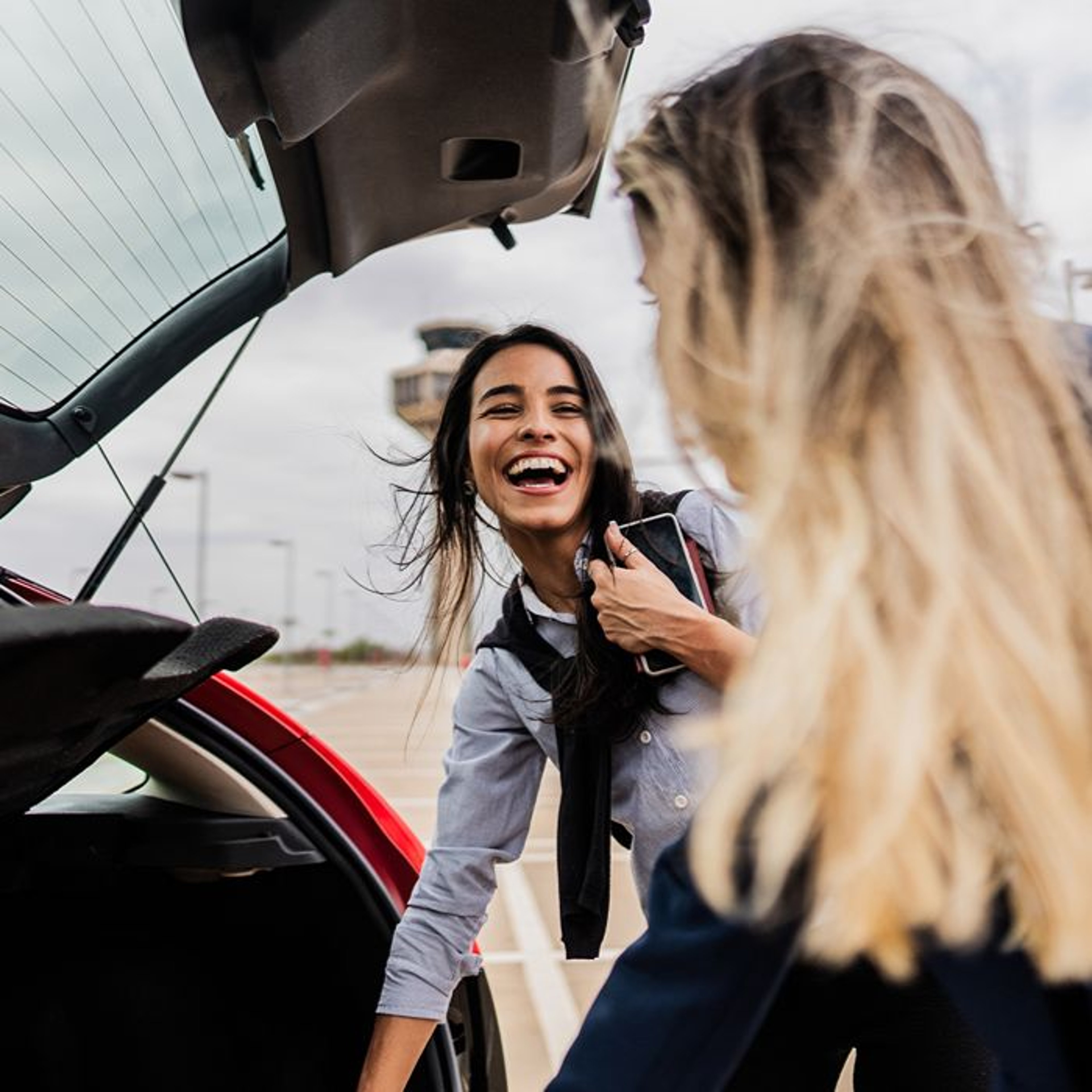 Two women smiling and interacting near the open trunk of a red car in an airport parking lot.