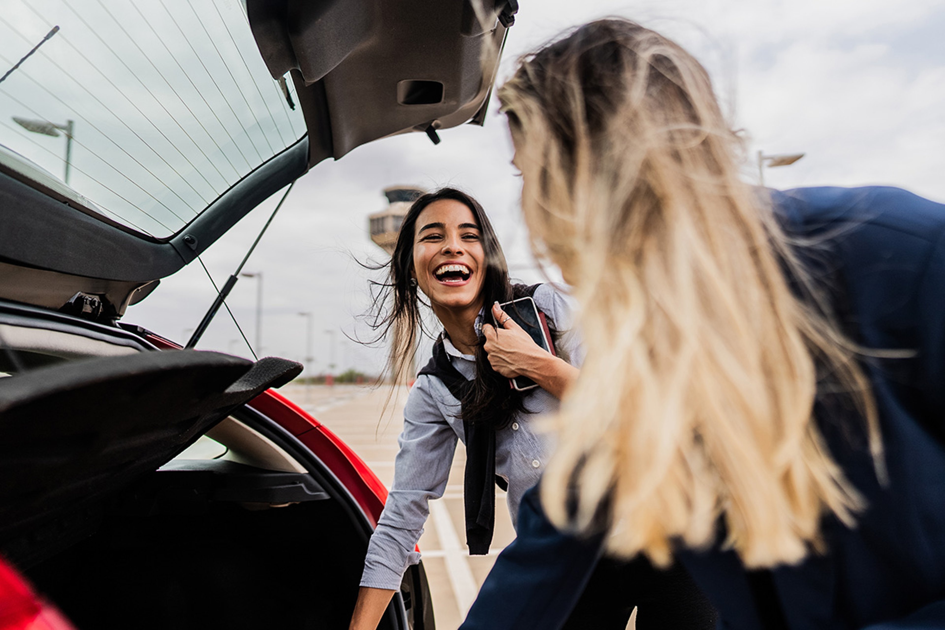 Two women engage by the open trunk of a red car in a parking lot, smiling and chatting.