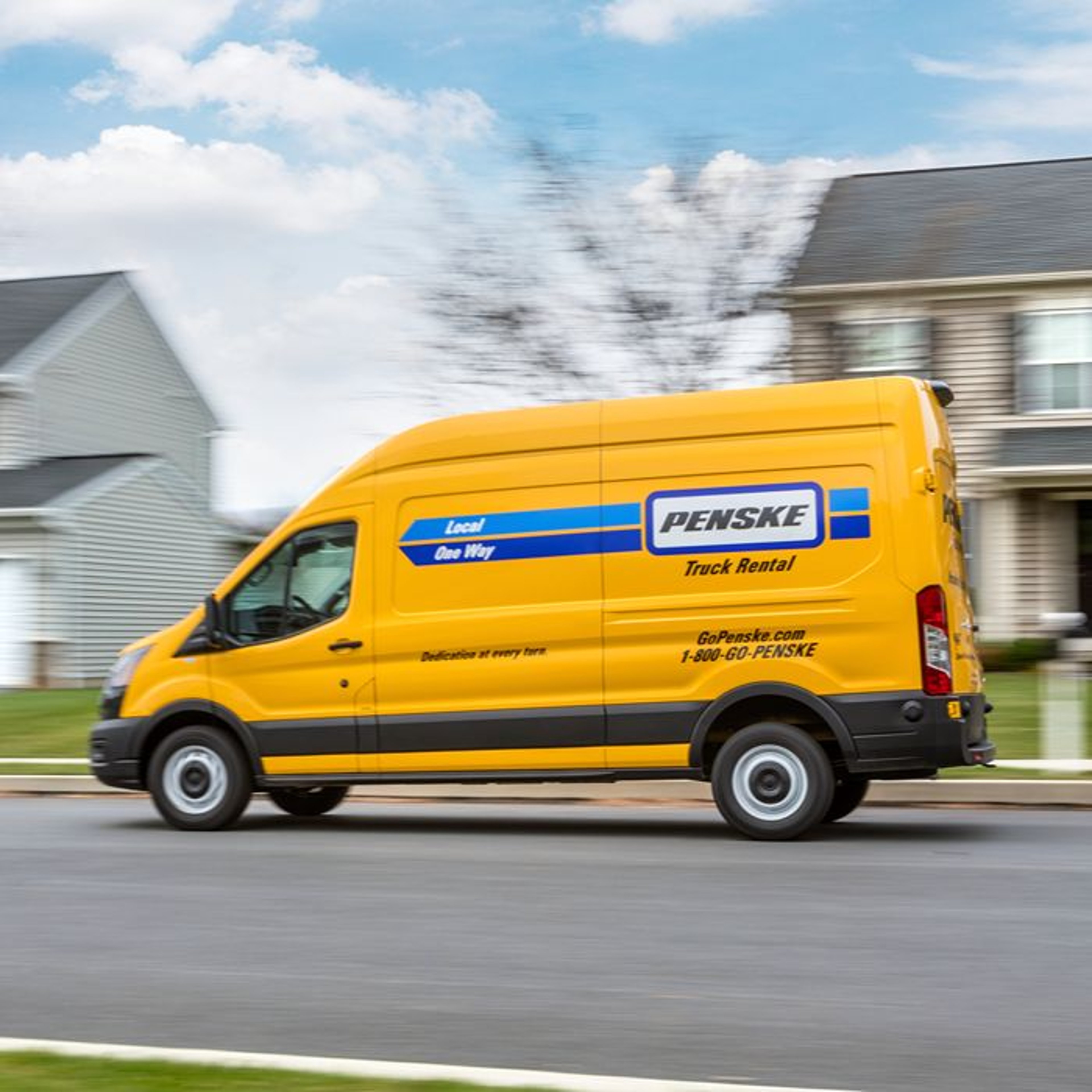A yellow Penske truck rental van is driving down a suburban street with blue skies and clouds in the background.